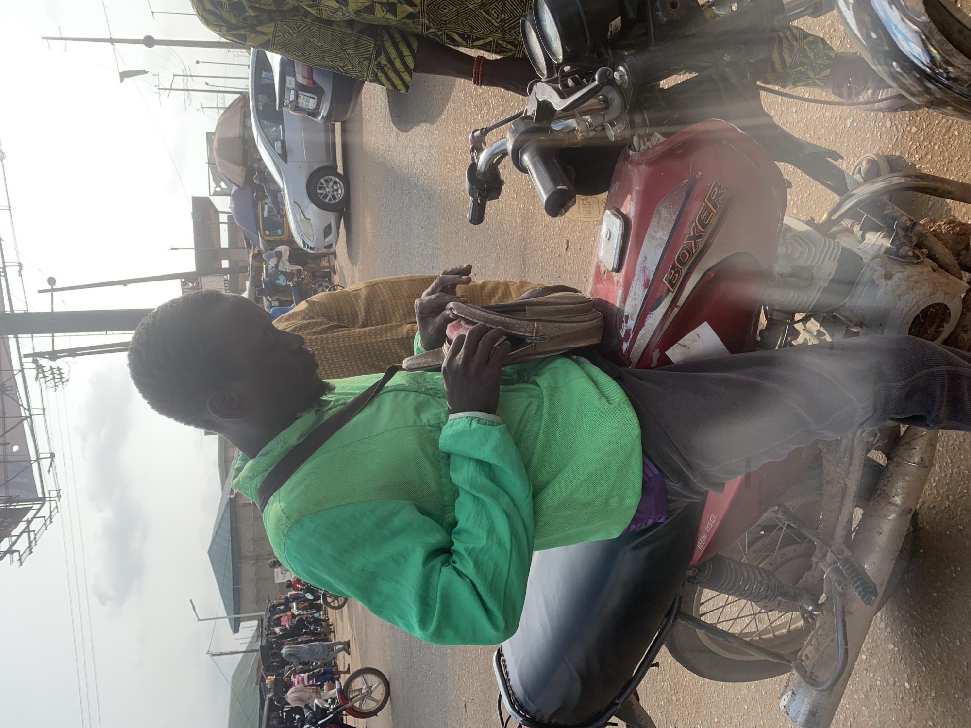A man in a green jacket and grey pants is sitting on a red motorcycle with "BOXER" written on its fuel tank. He is looking into a brown satchel that he is holding in his hands. Another man, wearing a yellow and brown patterned outfit, stands to his right, his hand resting on the motorcycle's handlebars. The motorcycle is parked on a dirt road, with other motorcycles and cars visible in the background. Several market stalls with green roofs are also present, along with power lines and street lights, suggesting an urban environment in Shagamu, Nigeria. The sky is overcast.