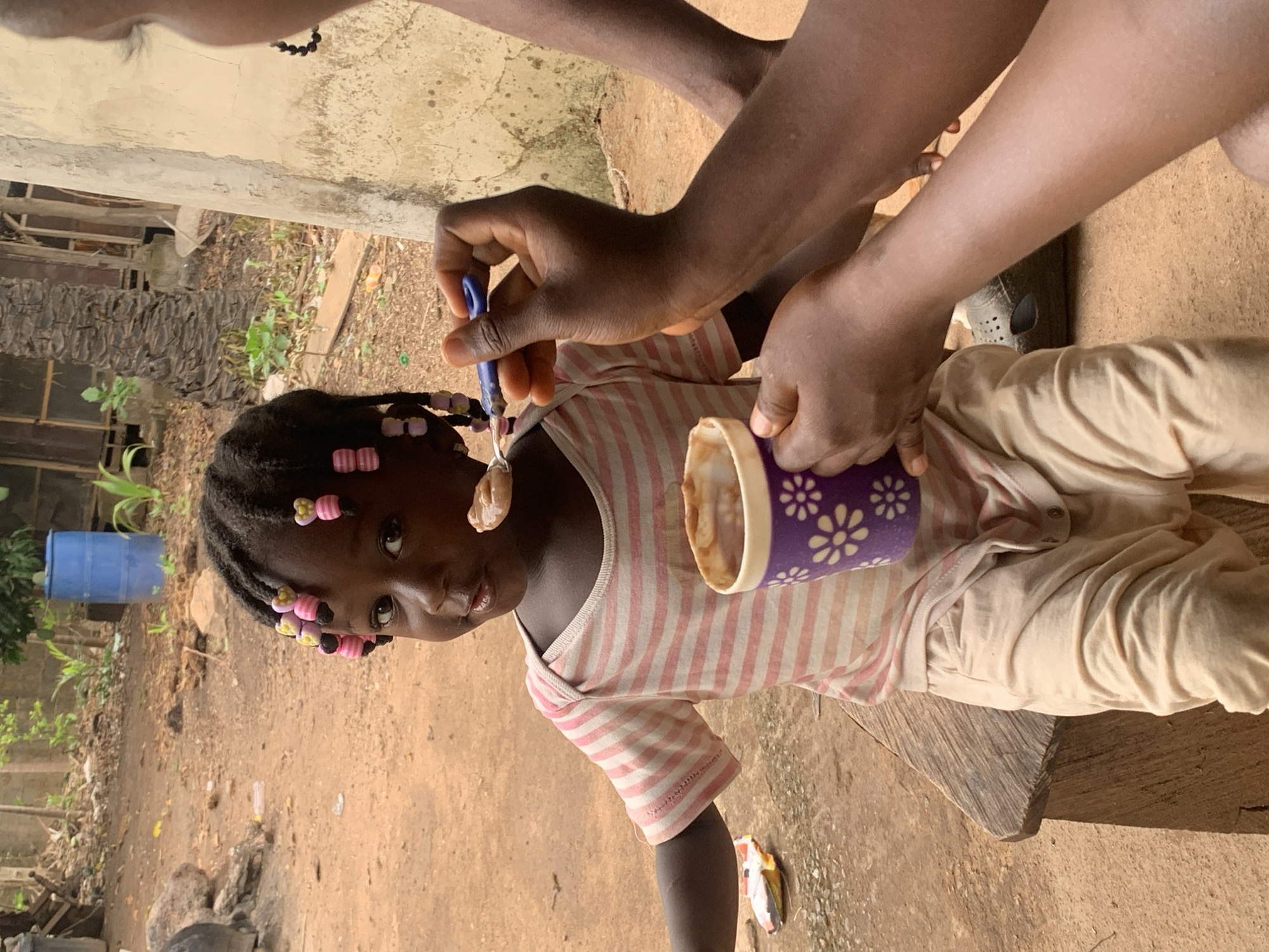 In Akwanga, Nigeria, an African child is being fed outdoors. She wears a striped shirt and has pink and yellow beads in her hair. An adult's hands are visible, one holding a spoon with food and the other holding a cup with a purple floral design. The child sits on a rough-hewn wooden bench in a dirt-covered yard. In the background are trees, a blue barrel, and a building wall.