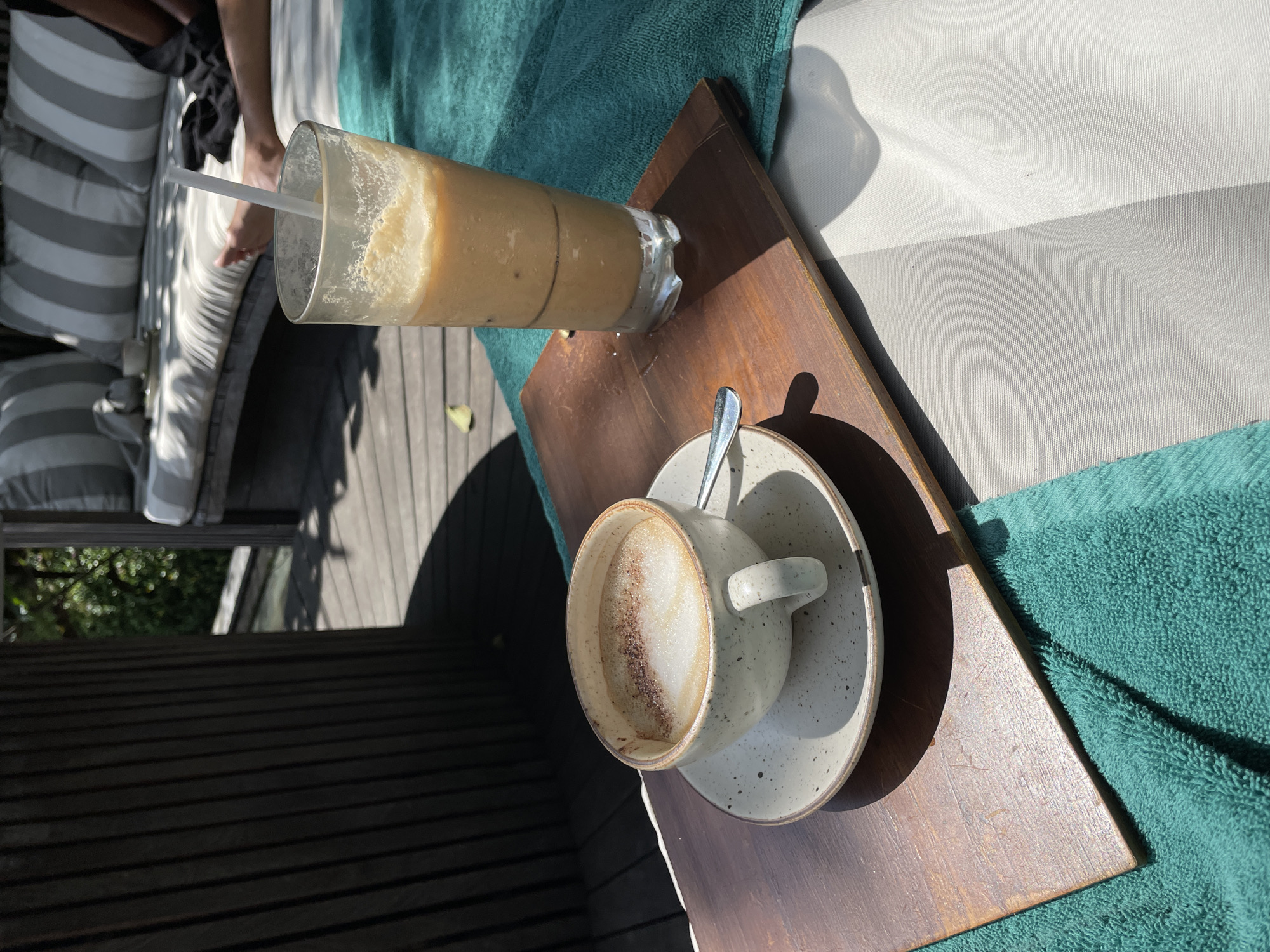 In Ubud District, Indonesia, a close-up shot shows a wooden tray with two drinks and a coffee cup on a turquoise towel. The tray sits on a lounge chair with a striped cushion. In the background, a wooden structure casts a shadow, and lush greenery is visible. The scene suggests a relaxing outdoor setting, possibly at a resort or cafe, with one person's legs visible on the lounge chair.