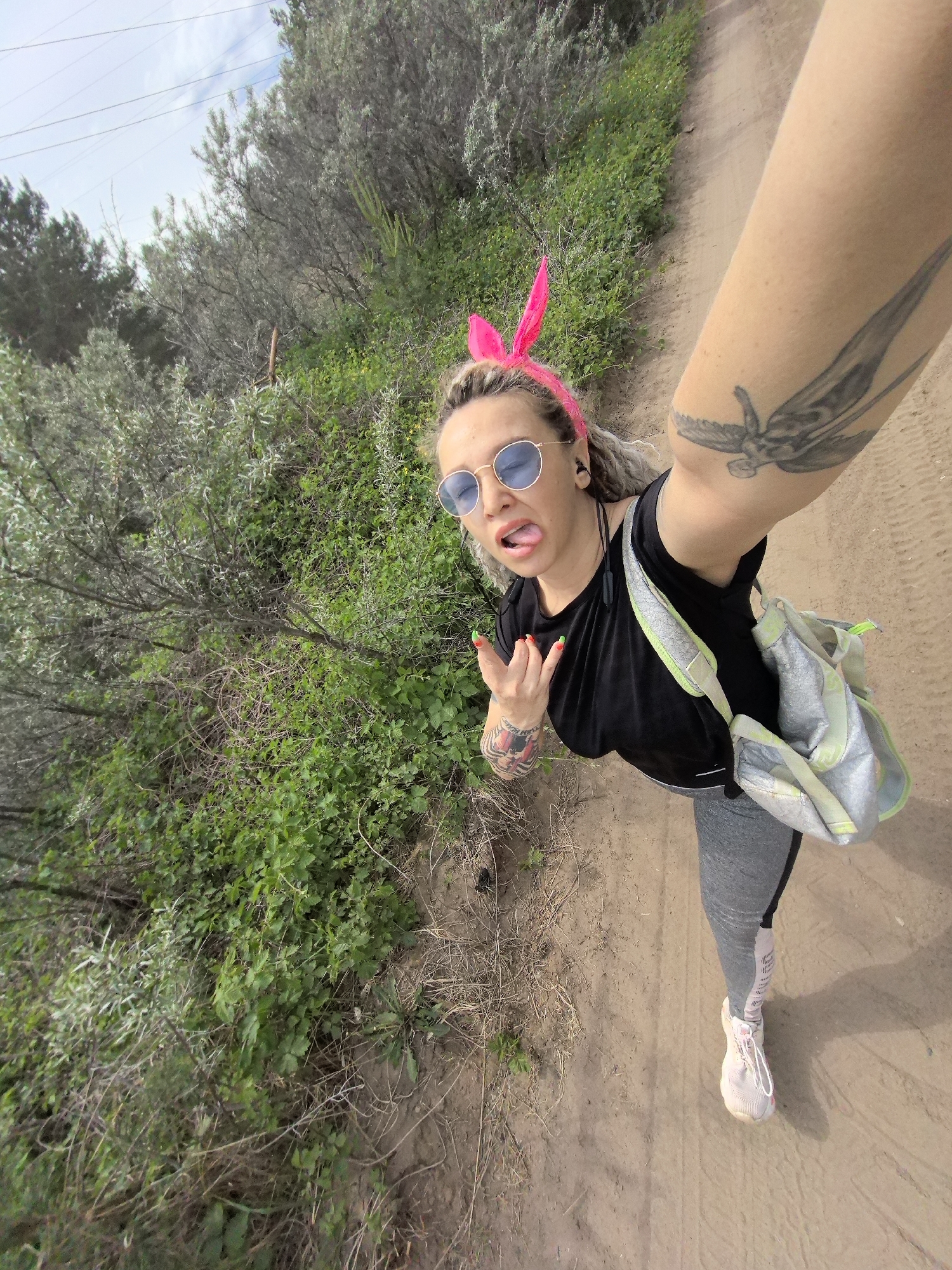A woman takes a selfie on a dirt path in a natural setting. She wears pink bunny ears, round blue sunglasses, a black t-shirt, gray leggings, and pink sneakers. She has a gray backpack over her shoulder and tattoos on her arm and hand. She sticks out her tongue and makes a hand gesture. The path is surrounded by green vegetation, possibly shrubs and trees. Power lines are visible in the upper left corner. The location is Noviy, Russia.