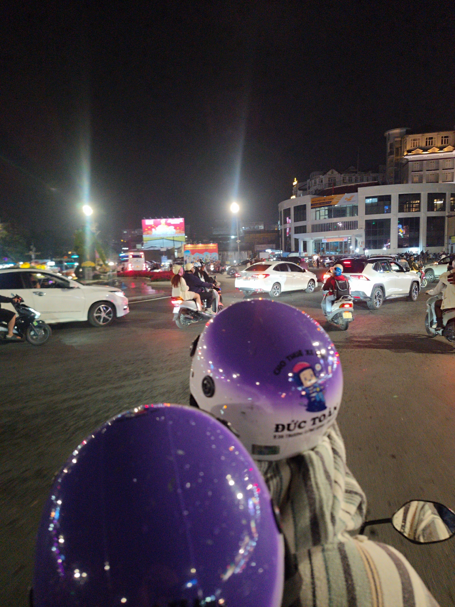 The image captures a vibrant night scene in an urban environment, likely in Da Lat, Vietnam, from the perspective of a person on a motorcycle. In the immediate foreground, two distinct purple motorcycle helmets are prominent. The closest helmet is partially visible on the bottom left, while a more complete one occupies the center-right, adorned with sparkly accents. This visible helmet features Vietnamese text, including "CƠ THỂ KỊP" (partially readable) and the name "ĐỨC TOÀN," alongside a small cartoon character. The person wearing this helmet is seen wearing a striped jacket or top. A motorcycle mirror is also visible on the far right.
The midground depicts a busy city street with multiple lanes, illuminated by numerous artificial lights. A mix of vehicles, primarily motorcycles and scooters, along with several cars (including a white sedan and a white SUV), are navigating or stopped in traffic. People are seen riding the motorcycles, some with passengers, indicating active night travel or commuting. Headlights, taillights, and streetlights create bright streaks and reflections on the dark road surface.
In the background, multi-story buildings line the street, many with illuminated windows and facades. A prominent, brightly lit billboard displays colorful imagery and partially discernible Vietnamese text, including "CÚC HỌA MI." Another building sign shows the word "VIETNAM." The sky above is dark, characteristic of nighttime. The overall atmosphere is that of a bustling urban night, with constant movement and artificial lights illuminating the scene.