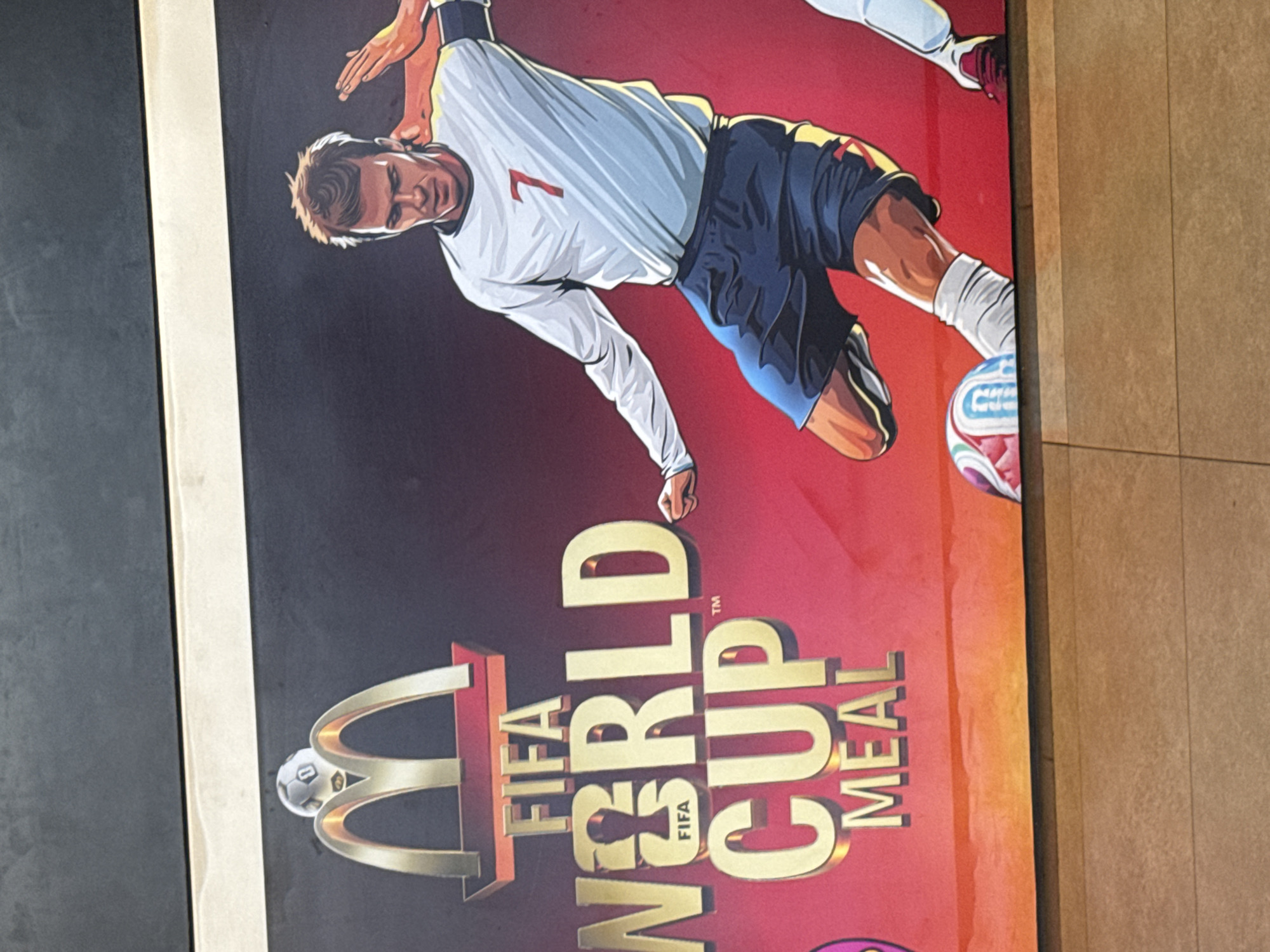 The image depicts a promotional poster featuring the FIFA World Cup Meal from McDonald's, set against a vibrant red background. The McDonald's logo, with golden arches and a football on top, is positioned in the upper left quadrant. To the right, a stylized illustration of a soccer player, identifiable as David Beckham due to his distinctive features and the number 7 on his jersey, is shown in action. He is wearing an England football kit and kicking a soccer ball. The text "FIFA WORLD CUP MEAL" is prominently displayed in large golden letters across the poster. A portion of another soccer player's leg and foot is visible on the right edge of the poster. The poster is likely displayed in a public area, potentially a fast-food restaurant or an outdoor advertising space in Kuta, Indonesia, given the context. The image is shot from a slightly upward angle, capturing the poster mounted on a wall with a wooden textured surface below it.