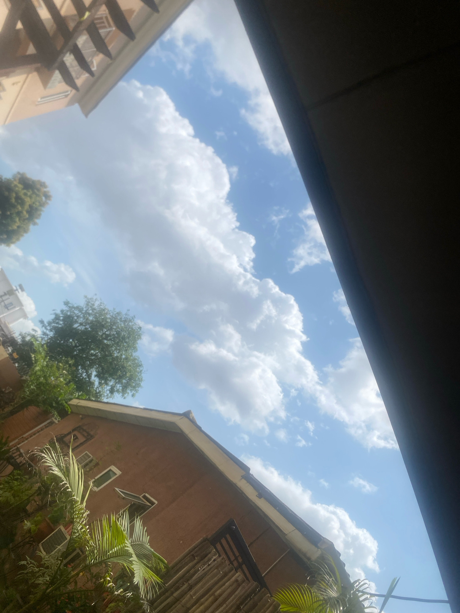 This is a photograph taken in Barden Yana, Nigeria, looking upwards towards a blue sky filled with fluffy white clouds. In the foreground, the roofline of a brick building with several windows is visible, along with lush green palm fronds and other foliage. To the left, part of another building with a wooden overhang can be seen. The right side of the image is dominated by a dark, shadowed area, possibly an overhang or a wall. The scene appears to be an outdoor residential or urban setting.