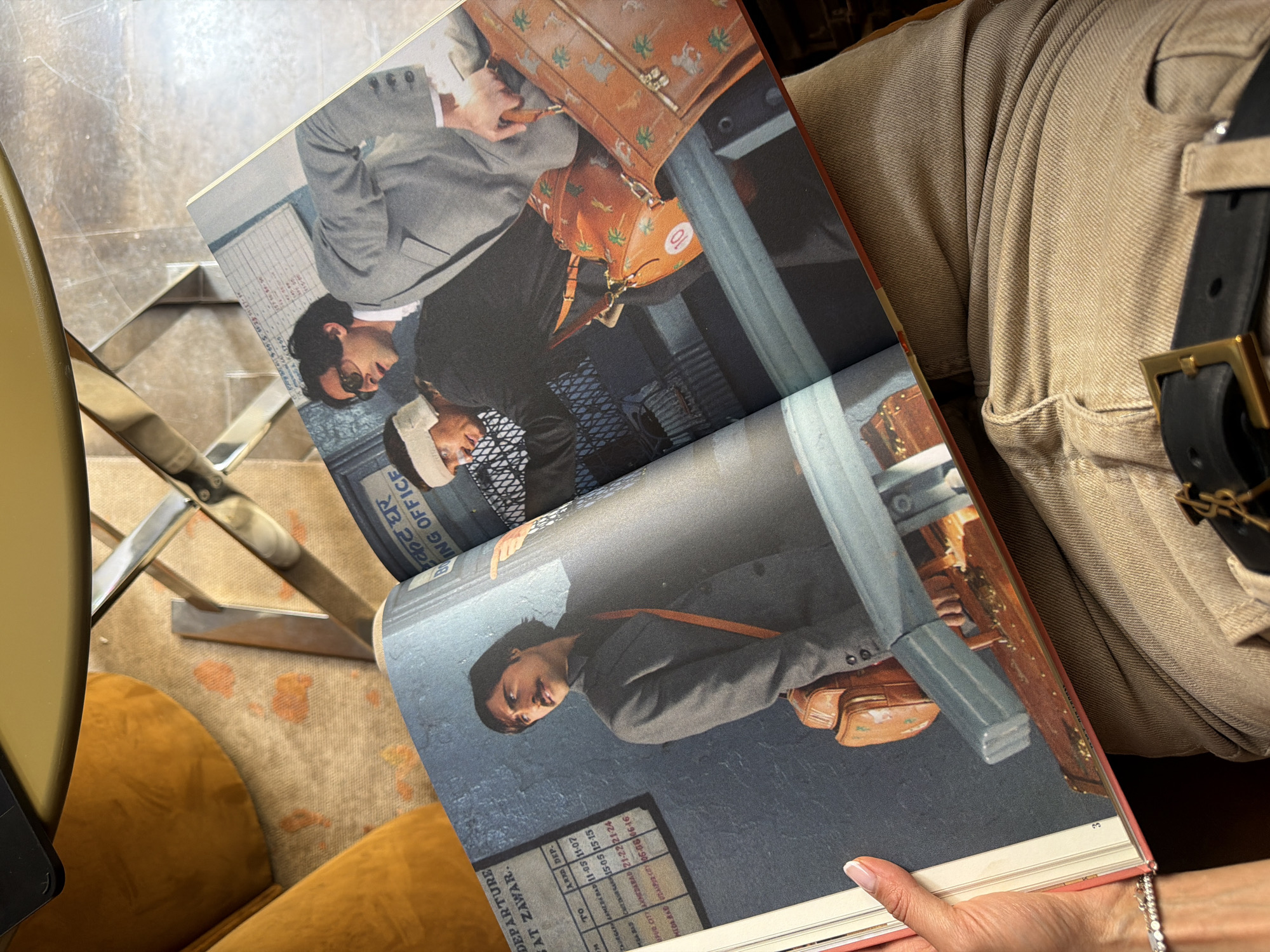 The image shows an open book held by a person wearing a bracelet. The book displays a scene with several men and luggage, possibly a film still or a photograph from a magazine. One man in the foreground wears a grey suit and has a bag slung over his shoulder. In the background, two other men are visible. One is wearing sunglasses and a grey suit, and another has a bandage on his head. The background elements suggest a transportation hub, possibly an airport or train station, with signs indicating departure information and the words "OFFICE". The overall setting appears to be indoors with a muted color palette. There is no information in the image to suggest a location in Saint Petersburg, Russia.
