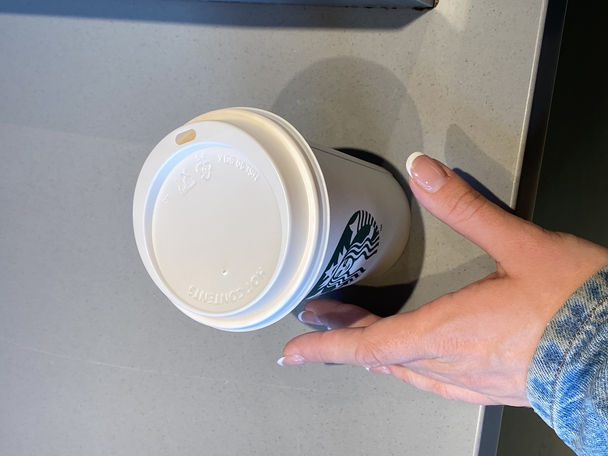 A top-down shot reveals a Starbucks cup with a white lid, its green logo partially visible, resting on a light grey countertop. A hand with manicured nails wearing a denim jacket extends toward the cup. The location is unspecified but assumed to be in Belgrade, Serbia. The scene suggests a momentary interaction or the placement of the coffee cup on the counter.