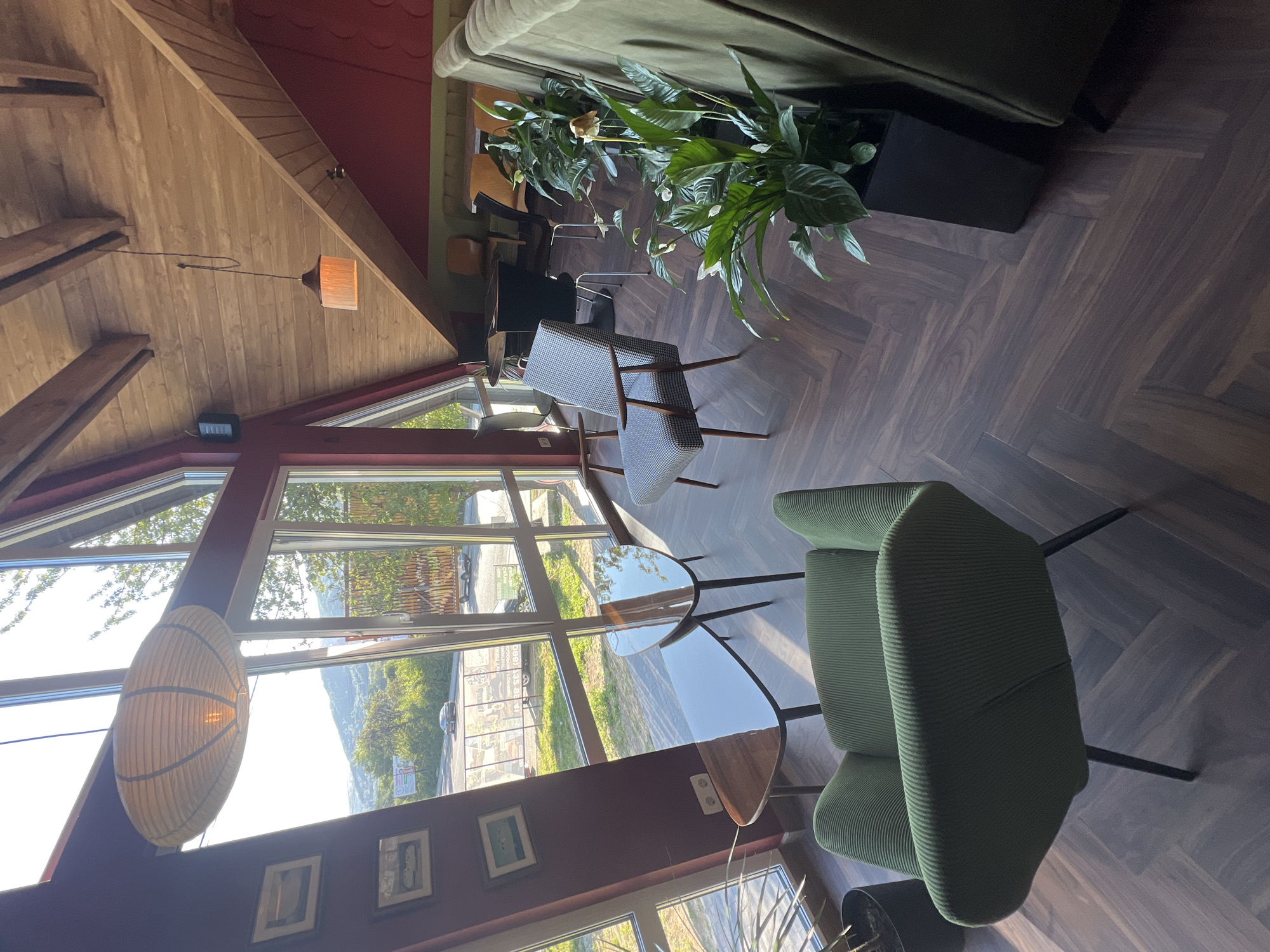 The image depicts the interior of a cafe with a sloped, wooden ceiling and large windows offering a view of a landscape outside. The cafe is furnished with several seating arrangements, including a green ribbed armchair in the foreground, a patterned armchair to the left, and a long, green sofa or booth along the right wall. There are also several small, triangular, wooden tables. In the background, a dark circular table with chairs is visible, suggesting a dining or meeting area. Several potted plants, including a peace lily, are placed throughout the space, adding greenery. Two pendant lights are visible, one large, oval, paper lantern-style light hanging near the windows and a smaller, cylindrical light hanging from the wooden ceiling. The flooring is a dark, wood-look herringbone pattern. Framed pictures hang on the maroon wall to the left. The overall ambiance is modern and relaxed, with a focus on natural materials and light. The specific location is identified as a cafe in Yaremche, Ukraine. No people are present in the image.