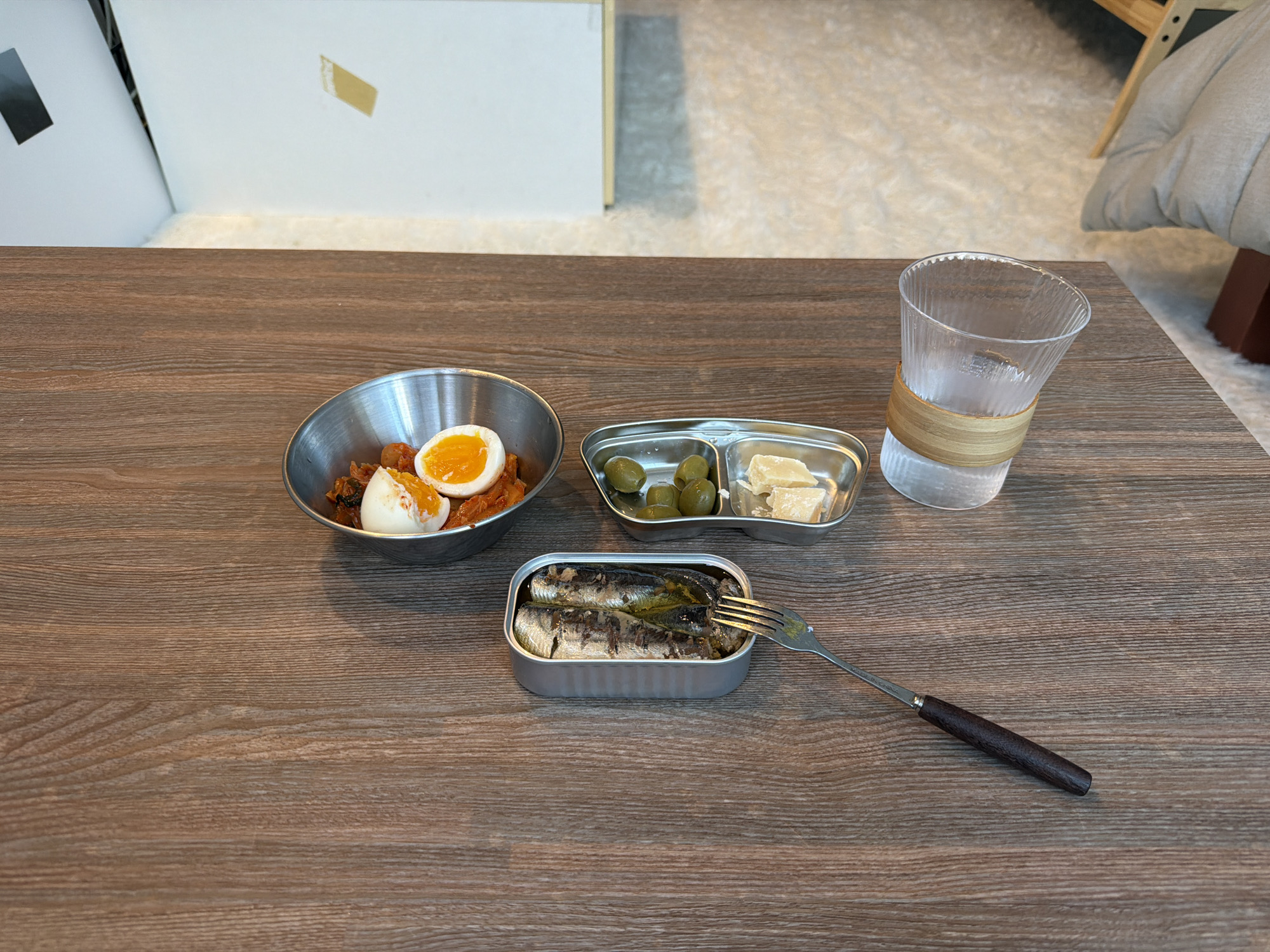 A high-angle, still-life photograph showcases a meal laid out on a wooden table, likely taken indoors. The scene includes a variety of food items and utensils arranged with no people visible.
To the left, a stainless steel bowl contains what appears to be kimchi and boiled eggs. Adjacent to this, a small, compartmentalized stainless steel tray holds green olives and what looks like chunks of cheese. Next to the tray is a glass of water with a woven band around its center. A can of sardines sits open, with a fork stuck into one of the fish.
The table's surface is a light brown wood with a visible grain pattern, suggesting it is made of real wood or a high-quality laminate. The background is blurred, but hints of a white wall or cabinet are visible, along with a glimpse of a patterned rug or floor.
Based on the style of food and dining presentation, the image could be taken at a meal in Seoul, South Korea. The lack of additional context prevents definitive confirmation.