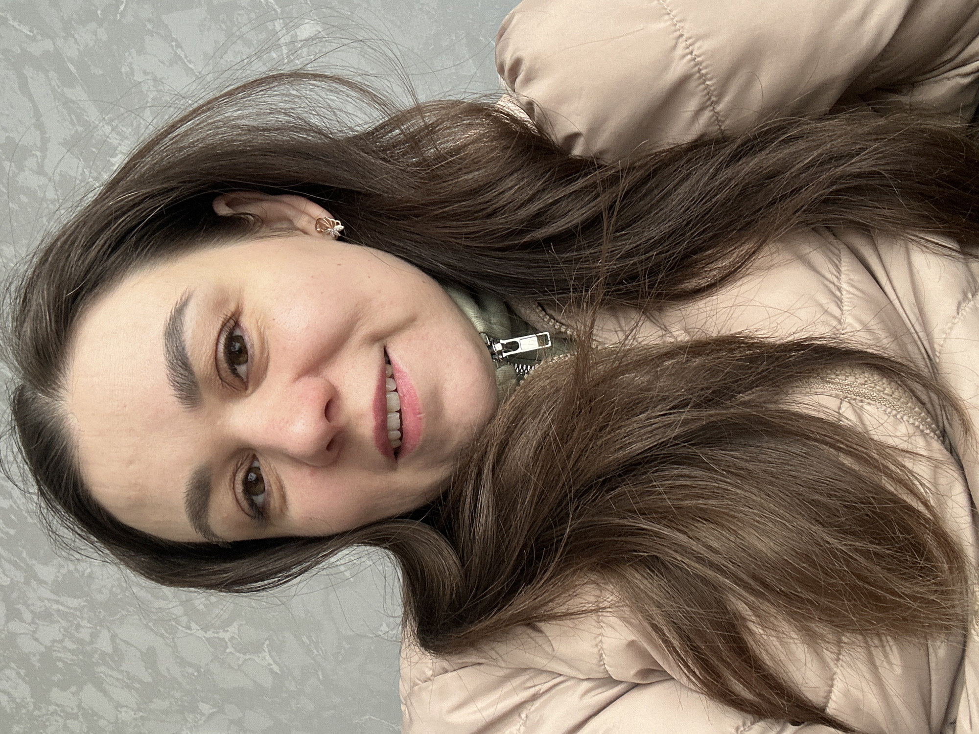 The image is a close-up portrait of a woman, oriented with her head tilted, suggesting she might be lying down or looking up. She has long, wavy dark brown hair cascading around her. Her face shows a gentle, open-mouthed smile, revealing her teeth, and her dark brown eyes are looking directly at the viewer. She is wearing light makeup with a hint of pink lipstick. A small, ornate earring is visible in her right ear. She is dressed in a light beige quilted puffer jacket, under which a green garment with a visible silver zipper pull can be seen. The background features a light grey wall with a subtle, textured pattern. The overall impression is one of friendliness and contentment as she poses for the photograph. No text is visible in the image. This scene takes place in Chaikovsky, Russia.