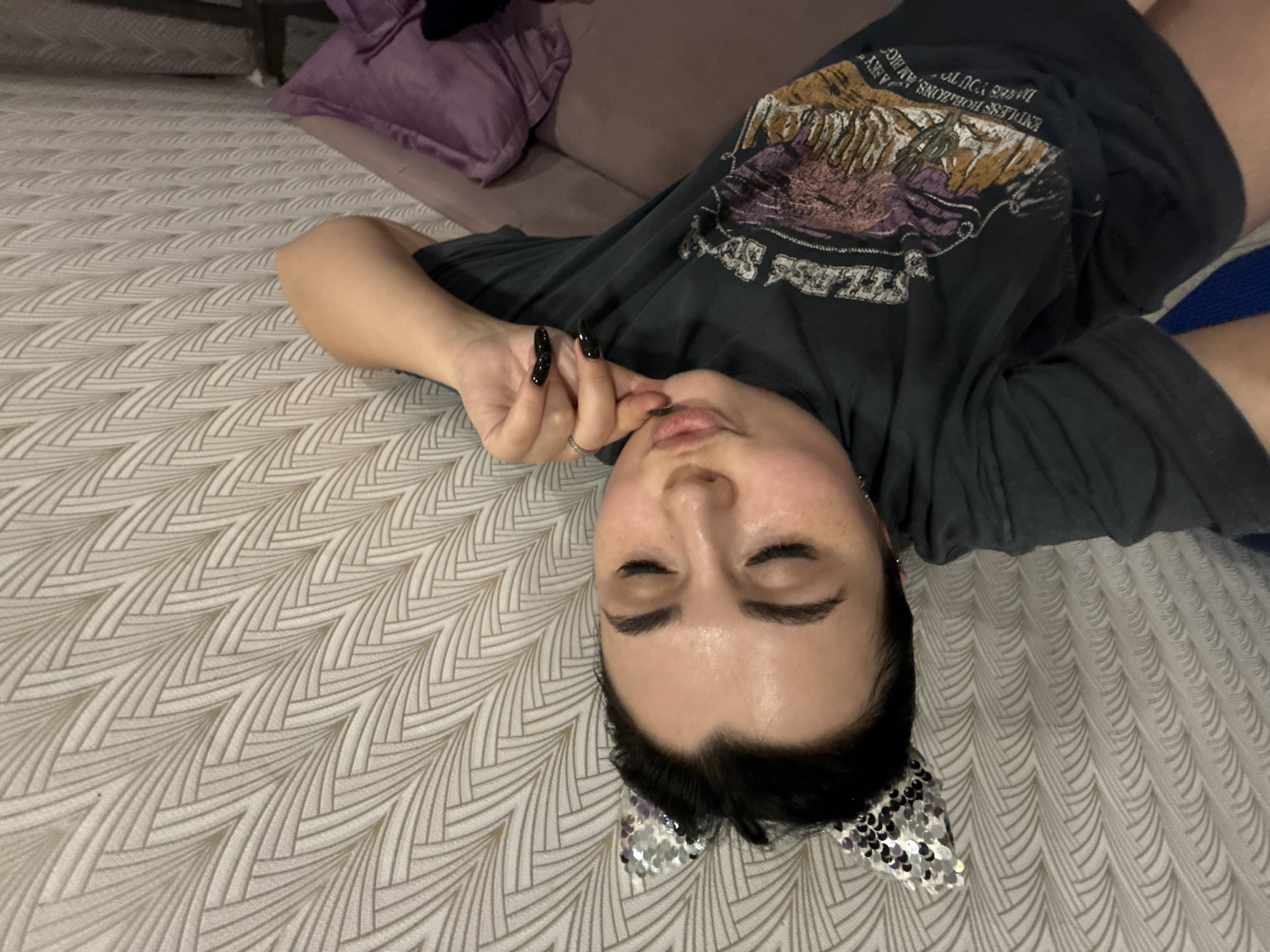 A woman is lying on a bed, wearing a black t-shirt with a graphic print and silver cat ears. She has black hair, fair skin, and long black nails. Her eyes are closed and she is holding her fingers near her lips. A purple pillow is visible in the upper left corner, and the bed has a patterned white and tan comforter. The location is not discernible from the image.