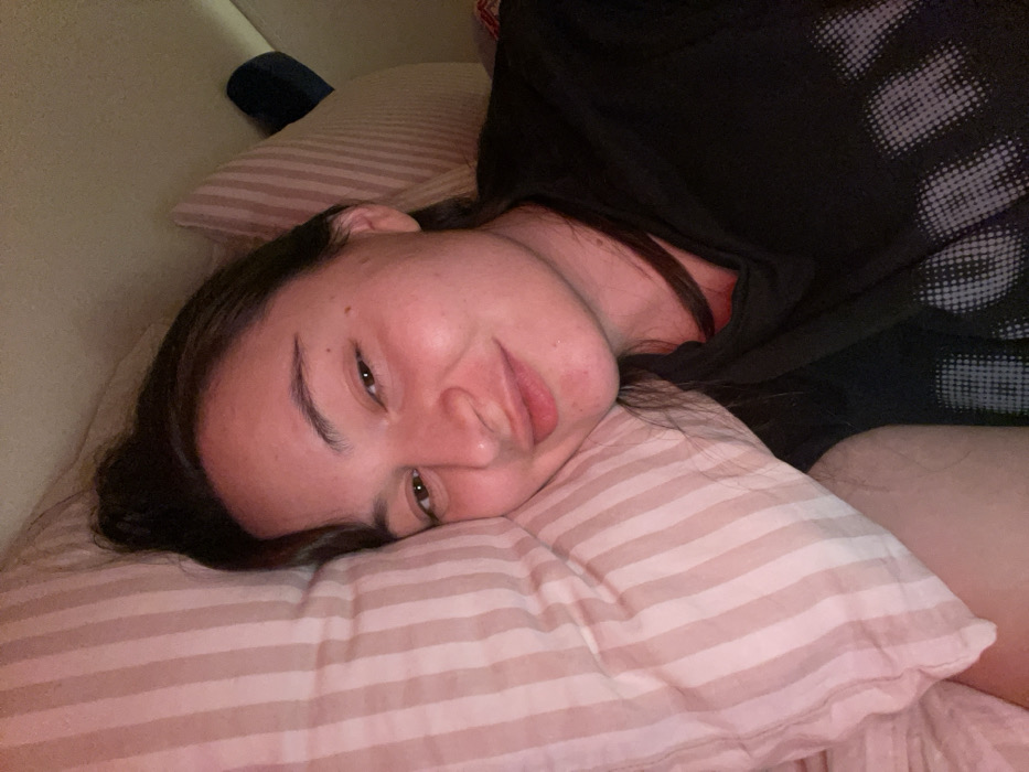 A person reclines on a striped pillow, their head resting comfortably. They are partially visible, wearing a dark shirt, and their dark hair contrasts with the light pink pillow. The person is facing forward, their gaze directed towards the camera with a slight smile. A wall is visible in the background.