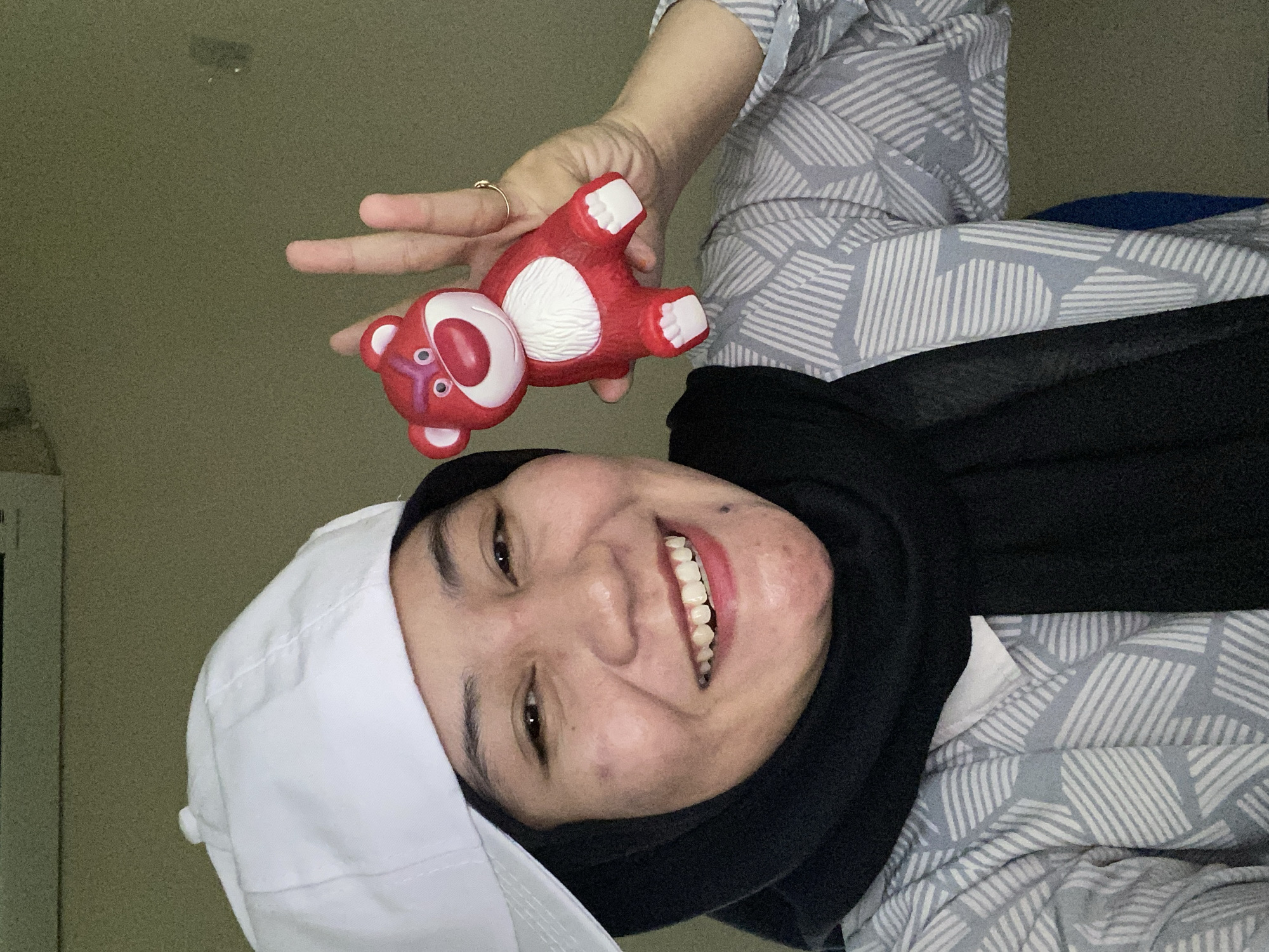 A woman wearing a white baseball cap and a black hijab smiles as she holds a red Lotso bear toy. The woman has brown eyes and is wearing a patterned grey shirt and a black scarf. The background appears to be an indoor environment with a muted green wall. The image was taken in Banjarbaru, Indonesia.