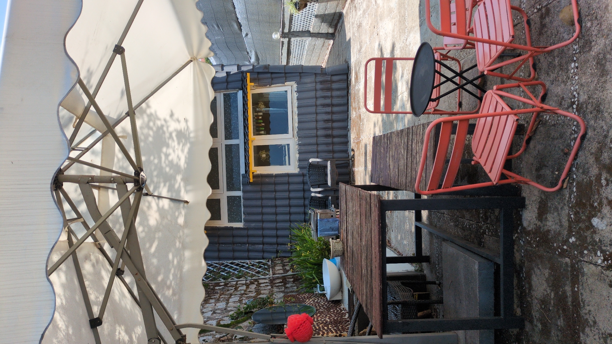 This outdoor scene depicts a patio or seating area with an umbrella, tables, and chairs next to a building. The building has a dark tiled exterior and windows with yellow trim. Garden decor and plants are present near the building's base. The seating area includes wooden tables with black metal frames and red metal chairs. A small black table is also present. The area appears to be paved with concrete. The location is Da Lat, Vietnam.