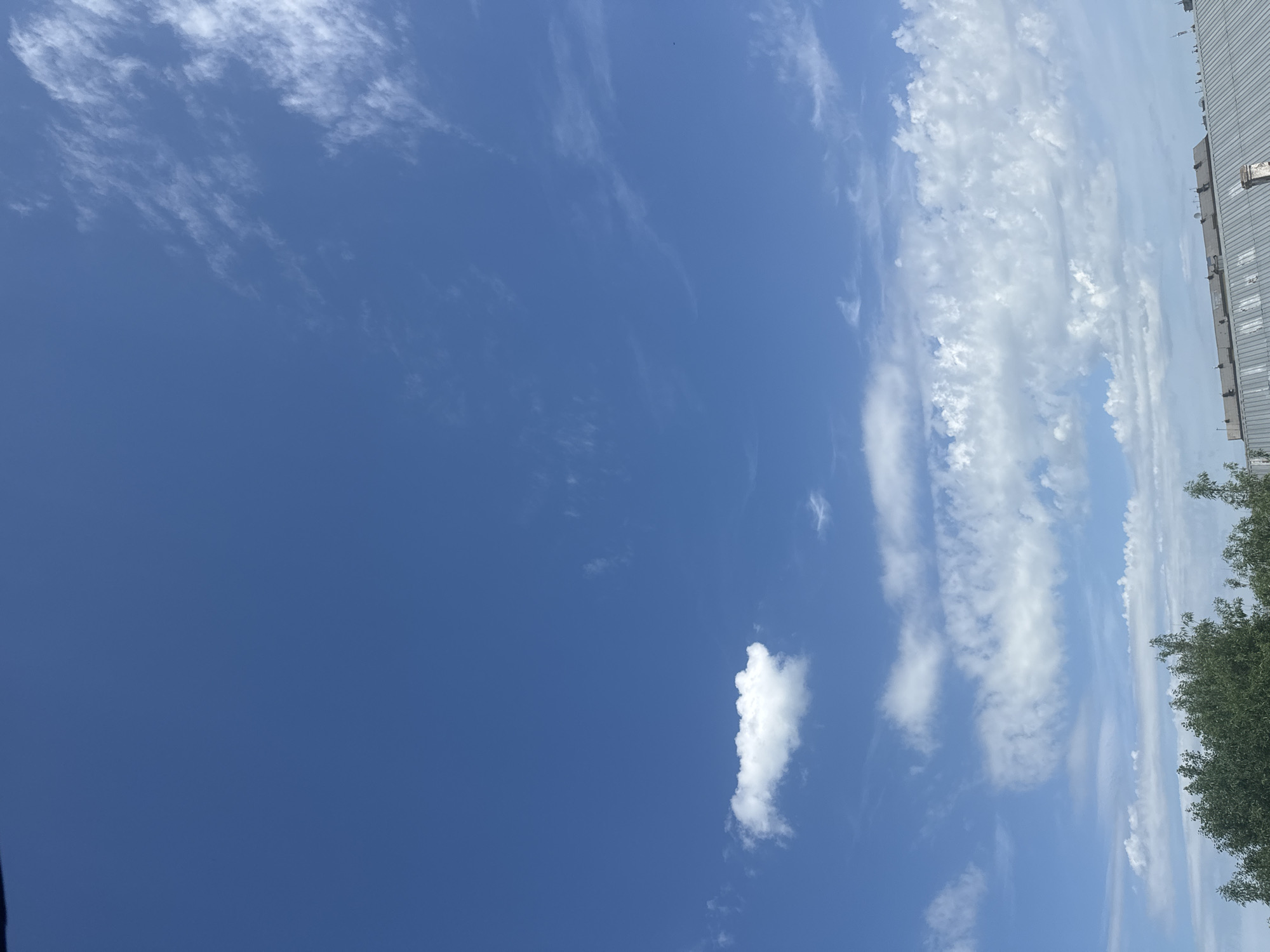 The image captures a wide expanse of a bright blue sky with scattered white clouds. In the lower portion of the frame, the tops of green trees and a metallic roof are visible, suggesting an urban or suburban setting. The scene appears to be a clear day in Aqköl, Kazakhstan, with no people or specific actions depicted. The focus is on the natural elements of the sky and vegetation, with hints of man-made structures at the bottom.