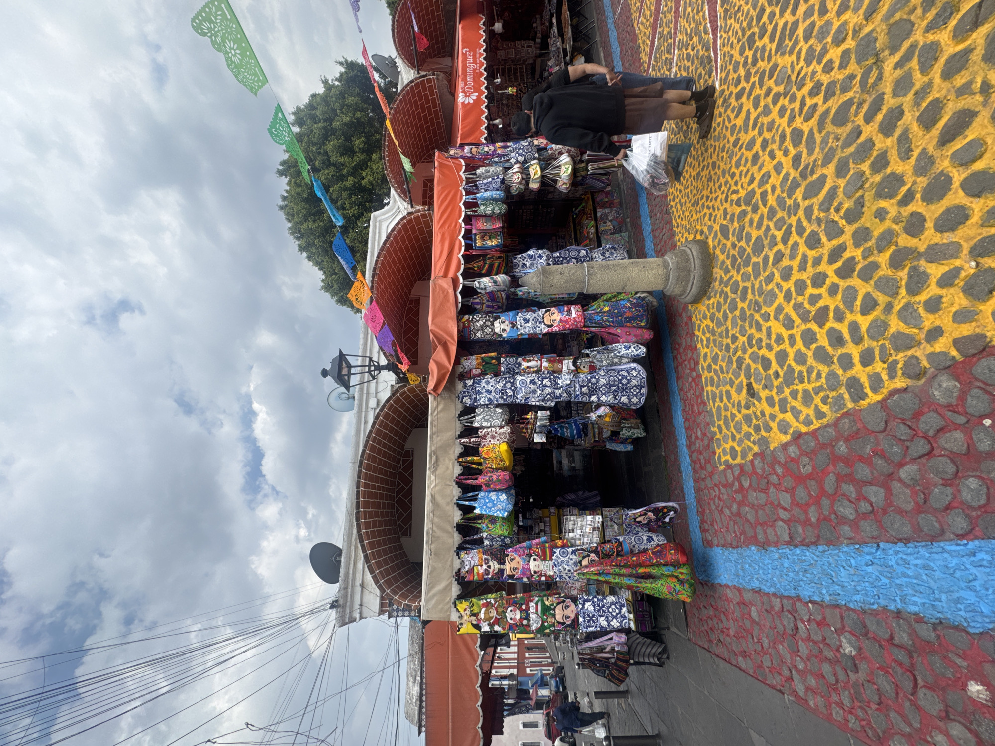 The image shows a souvenir stand in Puebla, Mexico. The stand is under an arched brick structure, with an orange awning labeled "Dominguez." Colorful paper banners are strung above. The stand displays numerous bags and souvenirs with Mexican designs, including images of Frida Kahlo. Two people are standing near the stand, appearing to be customers. The street is paved with stones painted in yellow, red, and blue patterns. Overhead are numerous electrical wires against a cloudy sky.