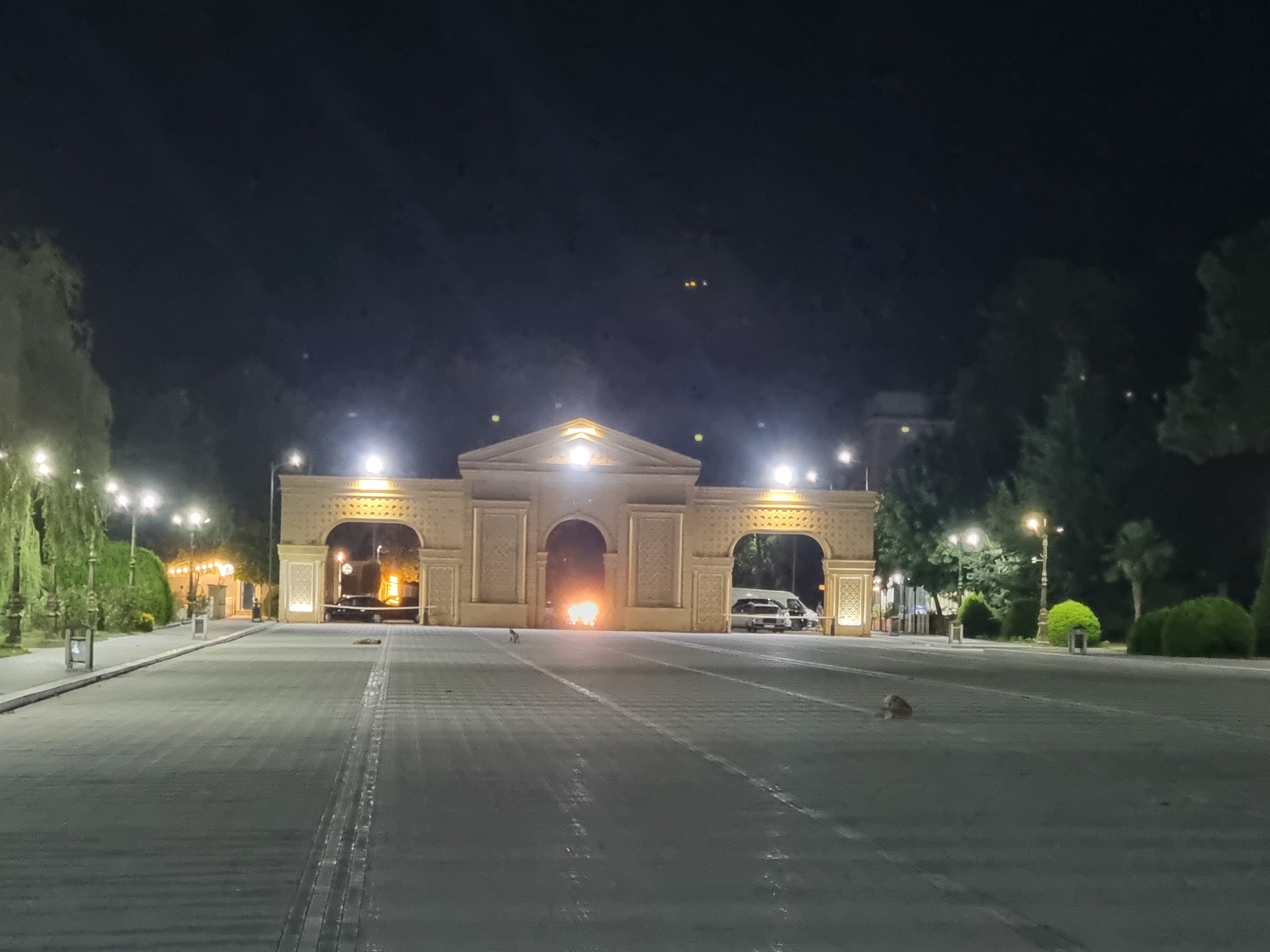 A wide paved plaza is illuminated at night by overhead streetlights and by lights emanating from a large, ornate archway in the distance. The archway, made of a light-colored stone, has intricate carvings and is brightly lit from within. Two vehicles, a white van and a darker vehicle, are parked beneath the archway. In the center of the archway, a small fire burns. A single dog is visible in the foreground, standing in the middle of the paved area. Trees and manicured bushes line the sides of the plaza and the area around the archway, also illuminated by streetlights. The sky is dark and clear.