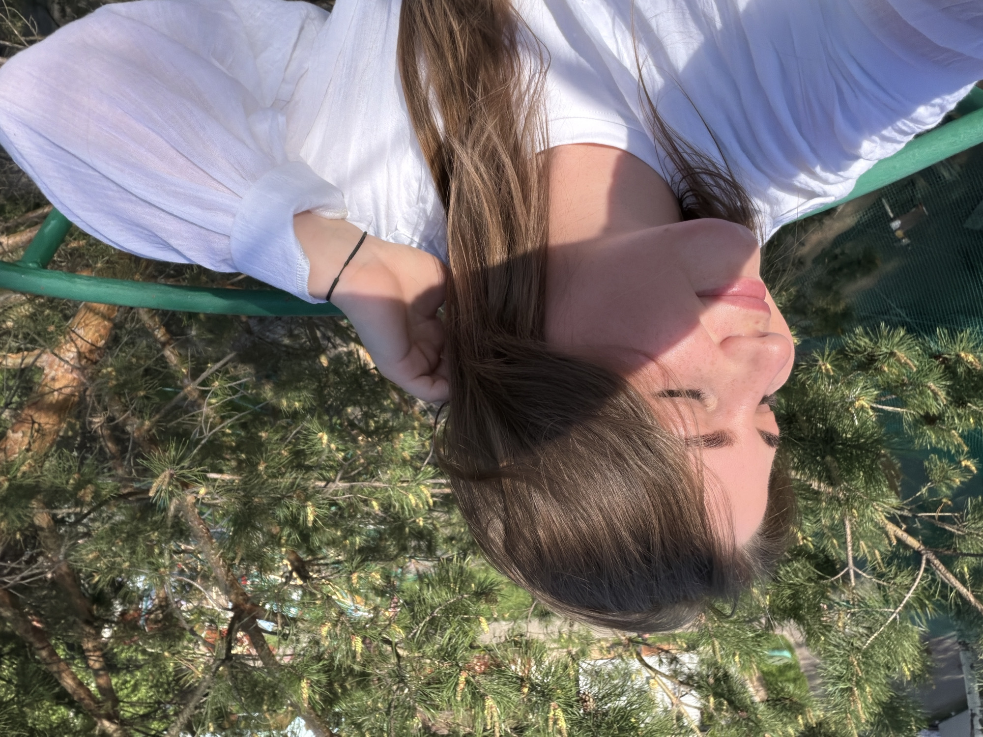 A woman with long brown hair is lying on a green bench, wearing a white blouse. She is outside, and evergreen trees surround the bench. The picture was taken in Chaikovsky, Russia.