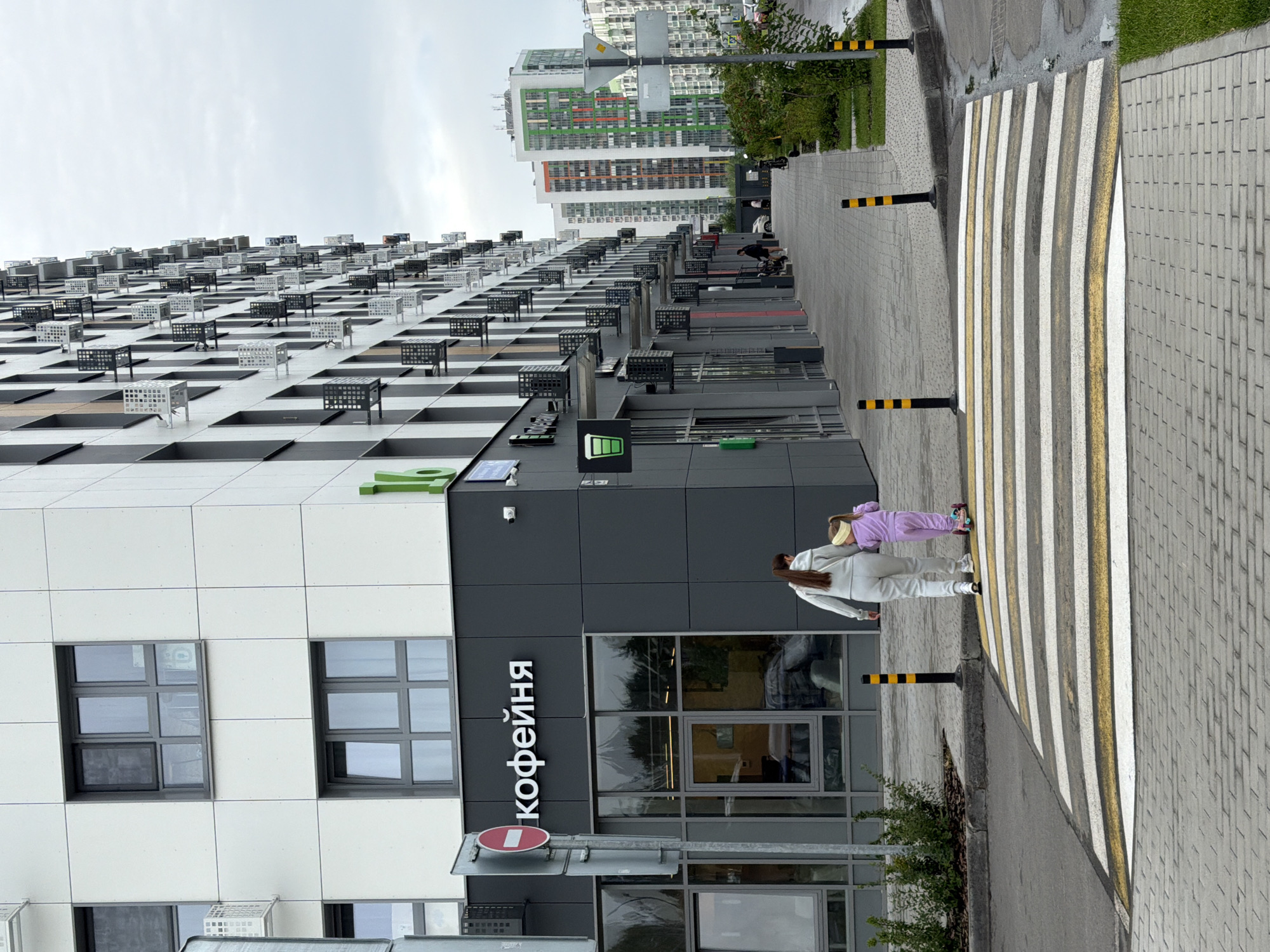 An overcast day in Kazan, Russia, features a modern residential building with multiple floors and a ground-level commercial space. A woman and a young child are walking across a pedestrian crossing. The woman is wearing a light-colored tracksuit, and the child, wearing a purple tracksuit and a yellow headband, is riding a scooter. The commercial space has a sign in Russian that reads "кофейня," indicating a coffee shop. The building is contemporary with many windows and balconies. The pedestrian walkway is paved with grey bricks. Yellow and black bollards mark the edge of the street. Lush green grass and bushes are visible along the side of the walkway. In the background, other similar residential buildings with colorful accents can be seen.