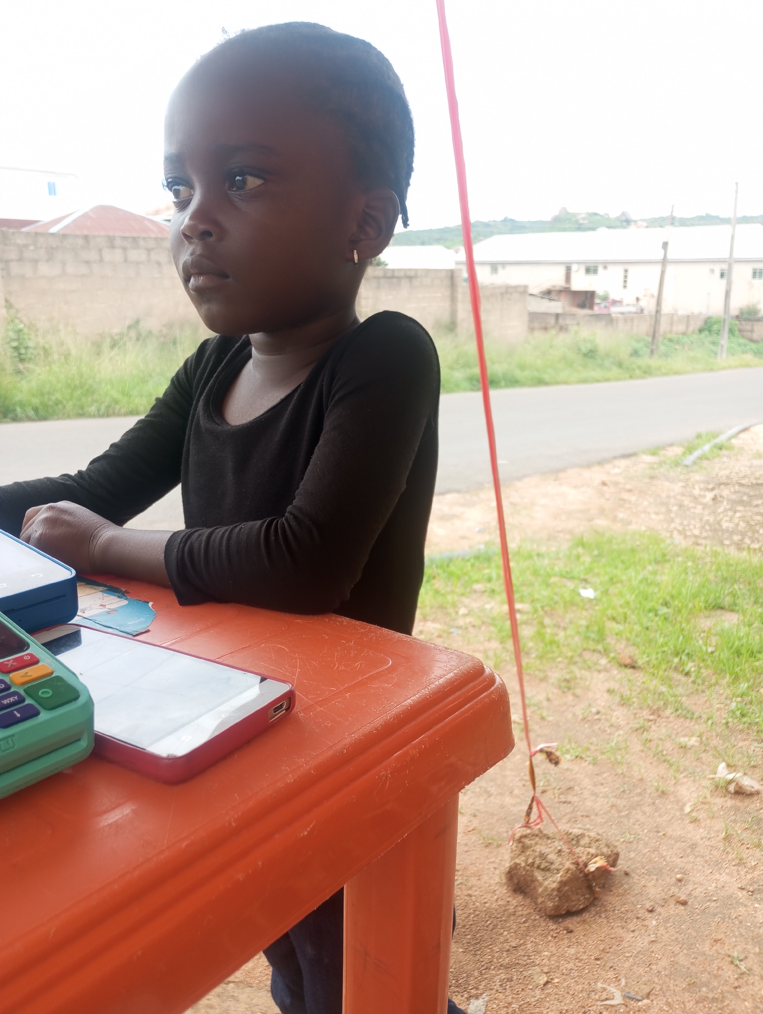 A young child, appearing to be of African descent, is seated at an orange plastic table outdoors, looking to their left. The child has dark hair, dark eyes, and is wearing a black long-sleeved shirt. On the table in front of the child are several items, including a light blue device, a red-cased smartphone, and a green electronic device with buttons and a screen. The background shows a road, green grass, a brick wall, and buildings, suggesting an urban or suburban environment. A red string is visible, tied to a rock on the ground. The lighting and overall scene indicate it is daytime. The image appears to have been taken in Jos, Nigeria, as indicated by the context provided.