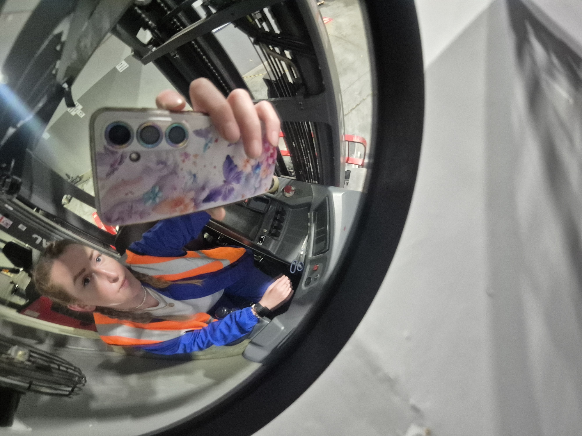 A person wearing a blue jumpsuit and an orange safety vest is sitting in a forklift, taking a selfie with a phone that has a butterfly-patterned case. The reflection in the curved glass of the forklift shows the person's face, the phone, and the interior of the forklift cab, including the steering wheel and control panel. The image is taken from a fisheye perspective, distorting the scene. The location is not specified in Chaikovsky, Russia.