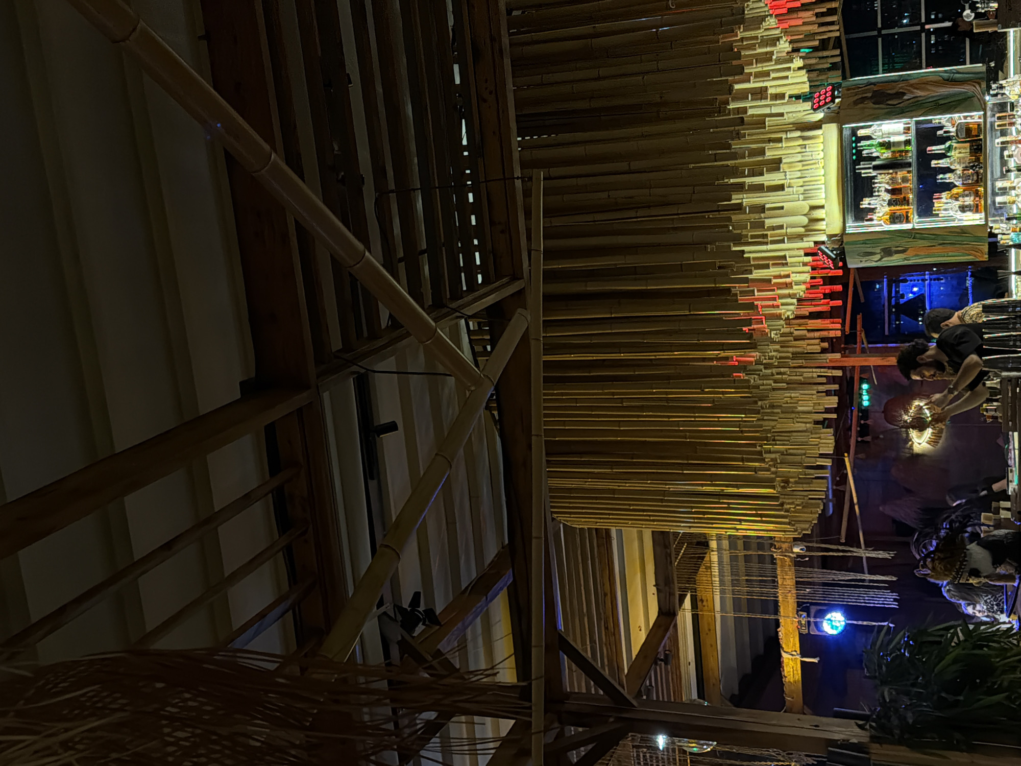 This indoor shot shows a bar/restaurant setting, likely in Yerevan, Armenia. The image is taken from a low angle, emphasizing the complex interior architecture. A bamboo-like structure serves as both decoration and possibly part of the ceiling. In the background, a well-stocked bar is visible with shelves of liquor bottles under bright lights. Near the bar, there are people, possibly customers and bartenders, although their faces are not clearly discernible. Decorative lighting and some greenery suggest a specific aesthetic design. The angle and framing of the shot create a sense of depth and complexity in the scene.