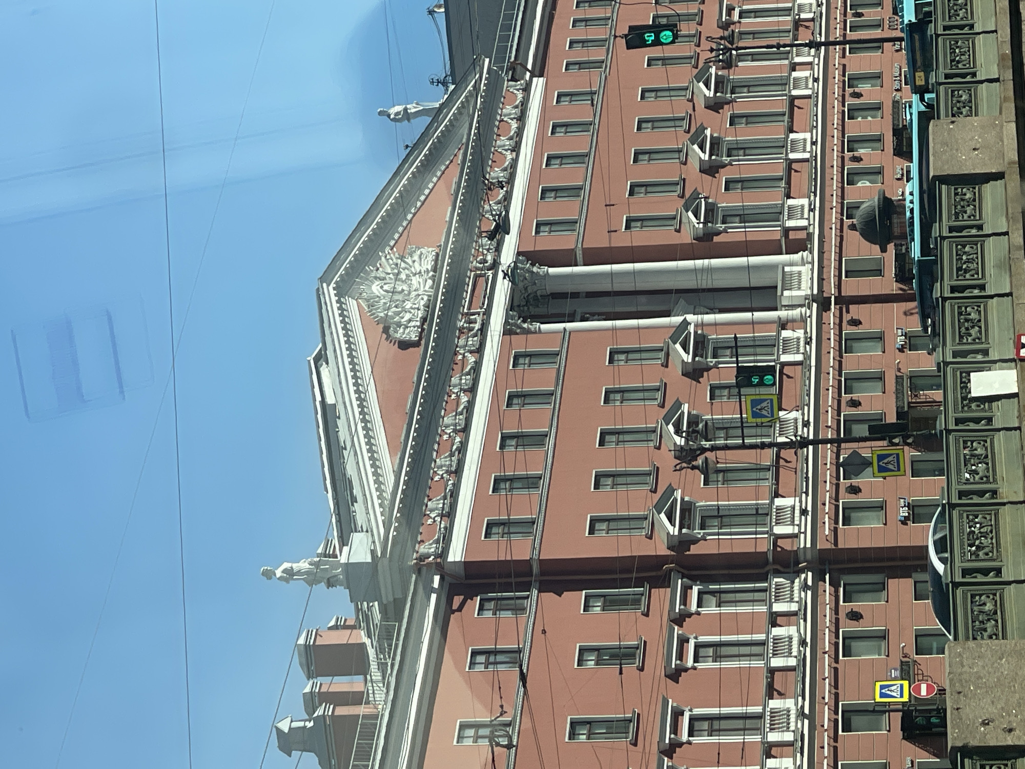 The image shows the exterior of a salmon-colored building with white architectural details, including statues and decorative elements on the facade. Windows with individual air conditioning units are visible on the building's face. A traffic light is seen, and a pedestrian crossing sign is attached to a light pole in front of the building. The photograph was likely taken in Saint Petersburg, Russia.