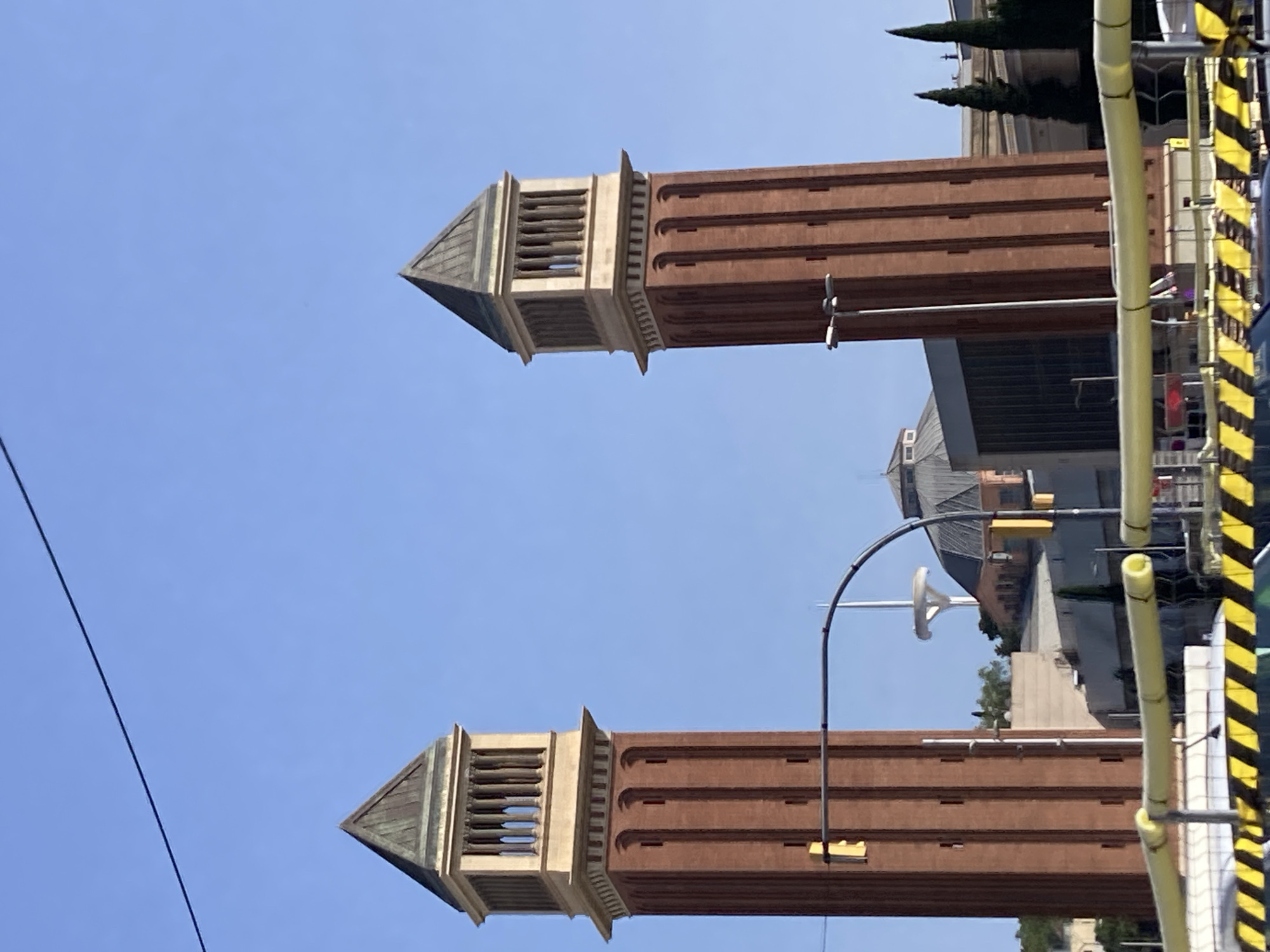 The image shows a portion of the Venetian Towers in Barcelona, Spain, against a clear blue sky. The towers are characterized by their reddish-brown brick construction and distinctive pyramidal roofs with slatted openings. One tower is positioned in the upper portion of the frame, and another is in the lower portion, suggesting a symmetrical or repeated architectural design. The towers are oriented horizontally in the image, indicating the photograph was taken with the camera rotated.
In the background, behind the towers, a building with a darker roof and lighter walls is visible. Trees, appearing to be cypress trees, are also present, adding a touch of greenery to the scene. A modern structure with yellow and black striped safety barriers and yellow cylindrical poles is partially visible on the right side of the frame, suggesting ongoing construction or maintenance work. The lighting indicates it is daytime, with bright sunlight. No people are visible in the frame. The image does not contain any visible text.