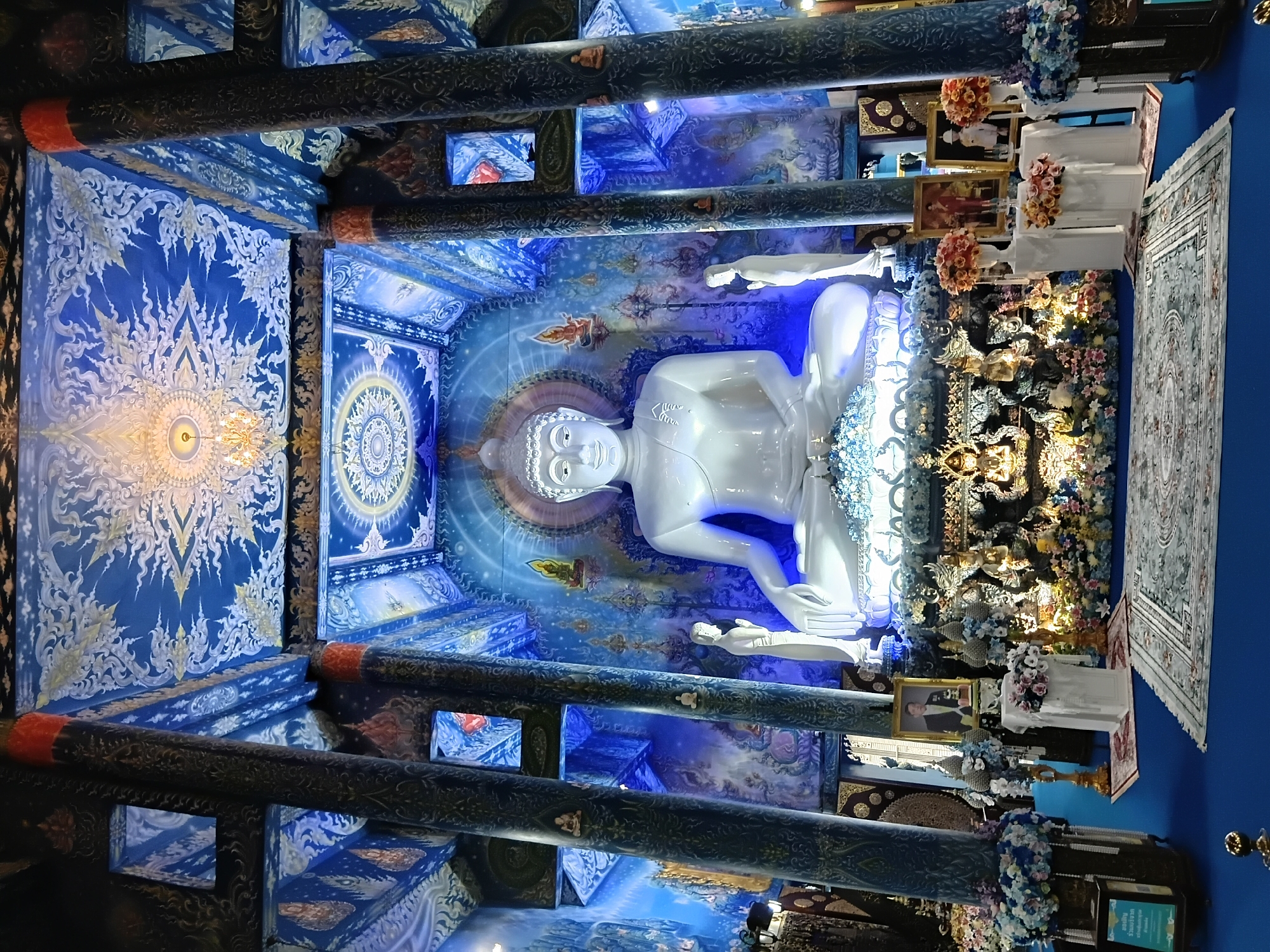 This image depicts the interior of the Wat Rong Suea Ten, also known as the Blue Temple, located in Chiang Rai City Municipality, Thailand. The central focus is a large, serene white Buddha statue seated on a raised platform adorned with intricate gold and floral decorations. Flanking the Buddha are two smaller, standing white figures. The surrounding walls and ceiling are elaborately decorated with vibrant blue murals, featuring celestial motifs and Buddhist imagery, accented by golden patterns. The architecture includes tall, dark blue pillars with ornate carvings, supporting a richly decorated ceiling with a chandelier. In the foreground, a patterned rug is laid out on a bright blue floor. Several framed portraits of men in formal attire are displayed on pedestals, surrounded by floral arrangements, suggesting a memorial or significant event. Artificial floral displays in shades of blue, white, and pink are placed throughout the space. The lighting appears to be a combination of ambient and focused illumination, creating a luminous and sacred atmosphere.