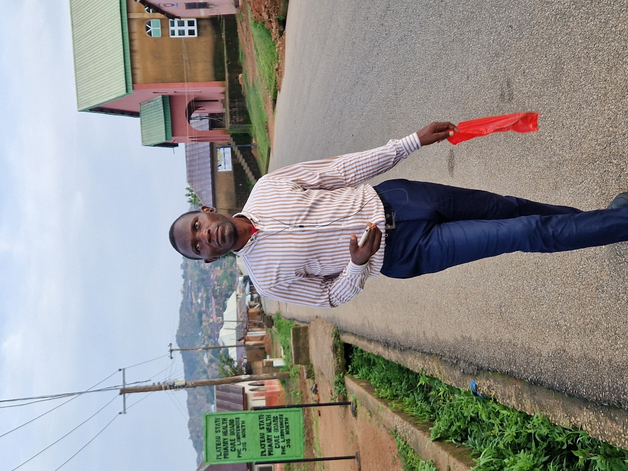 A Nigerian man walks down a paved road in a residential area in Kabong, Nigeria. He is wearing a striped button-down shirt and dark blue pants. He is holding a red plastic bag in his right hand and a smartphone in his left. The man is looking to his right. The road is lined with buildings on both sides, including a pink and green church. There is a drainage ditch filled with lush green vegetation to the left of the road. A green signpost with white lettering is visible on the left side of the road. The sky is overcast.