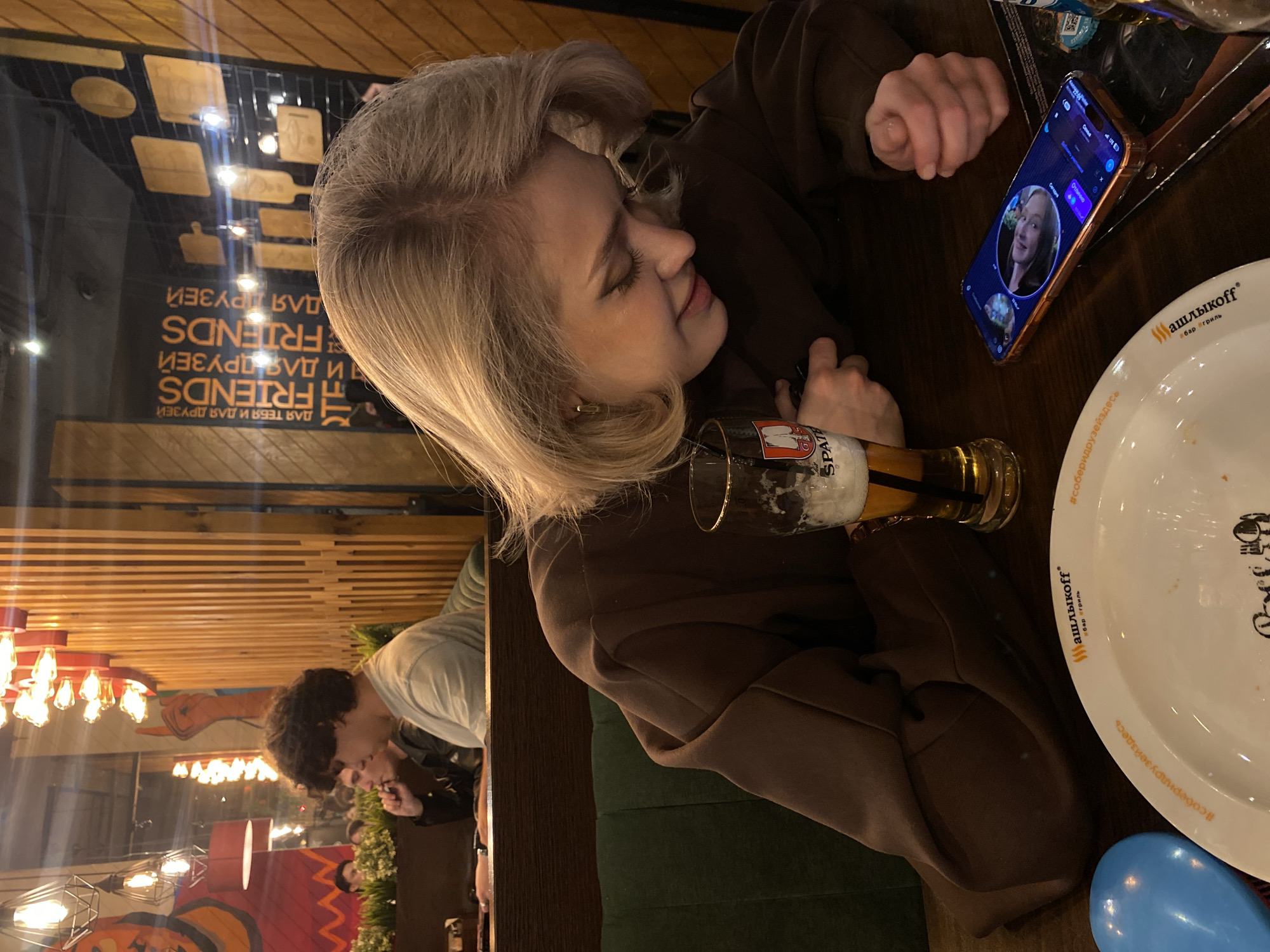 Here is a concise description of the image:
The image shows a woman with short blonde hair sitting at a table in a restaurant, with a glass of beer and a plate in front of her. She is looking at a cell phone with a person's face on the screen. The plate is marked with the brand name "Шашлыкоff", a bar and grill, suggesting a location in the city of Ufa, Russia, where this chain is present. The table also features wooden paneling and is reflecting the surroundings. Another person is visible sitting at another table in the background. The overall setting implies a casual dining experience with a social interaction via a mobile device.