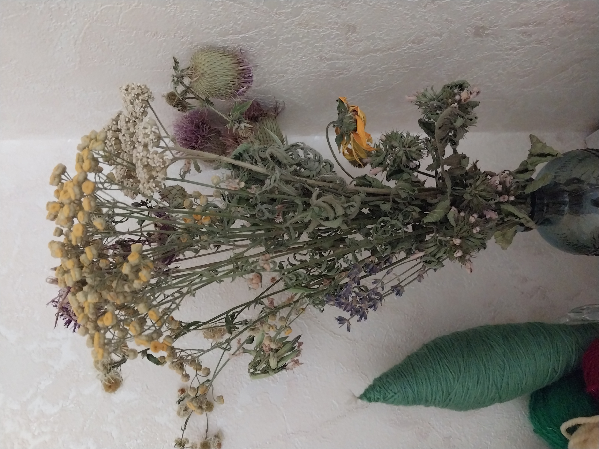 This is a close-up, eye-level, medium shot photograph of a dried floral arrangement in a dark glass vase. The bouquet is dense and varied, featuring a prominent cluster of small yellow flowers at the top, interspersed with finer white flowers and dried lavender sprigs. Several thistle-like seed pods in shades of green and purple are visible within the bouquet, adding texture. One dried yellow flower with drooping petals is prominently featured on the right side of the arrangement. The bouquet is set against a textured, light-colored wall. To the left of the vase, a tall, cone-shaped object wrapped in green yarn is partially visible, along with hints of other yarn balls in green and red. The overall impression is of a still life composition with natural elements. There are no people or animals present in the image. The scene does not provide any specific indicators to determine its location within the city of Dnipro, Ukraine.