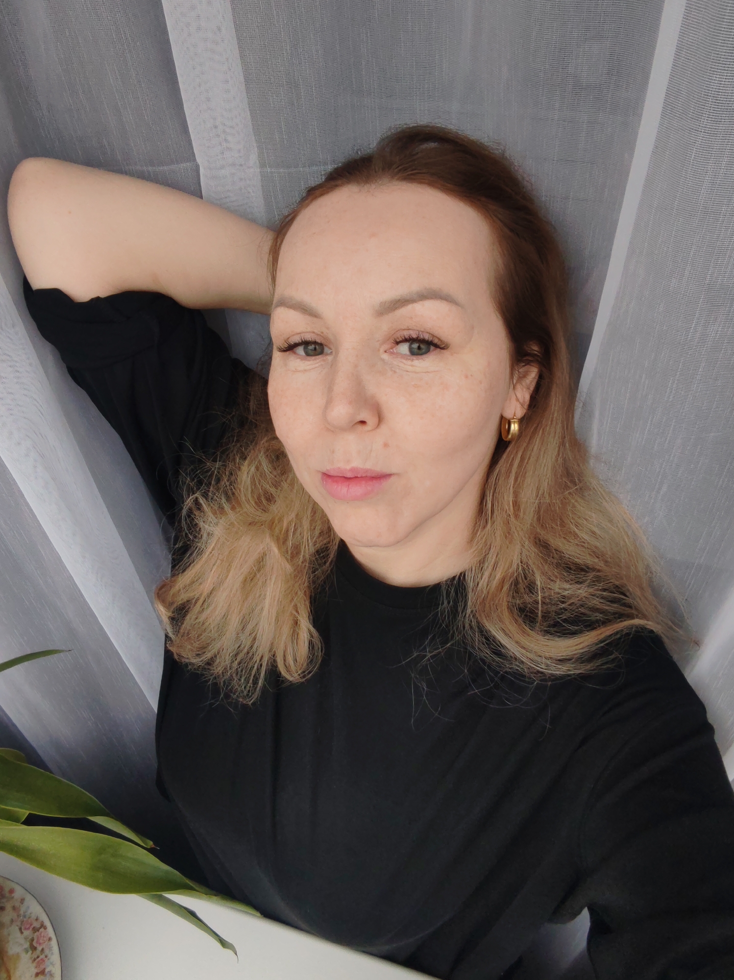 A fair-skinned woman with reddish-brown hair and freckles is posing for a selfie indoors, likely in a residential setting in Chaikovsky, Russia. She is wearing a black shirt and gold hoop earrings. Her left arm is raised above her head, resting on a light-colored or white sheer curtain. Part of a green houseplant is visible in the lower left corner, sitting on a white surface that may be a windowsill or table. A patterned plate or saucer is also partially visible near the plant. The background is dominated by the sheer white curtain, which appears to be part of a window or doorway covering. The lighting is soft and natural.