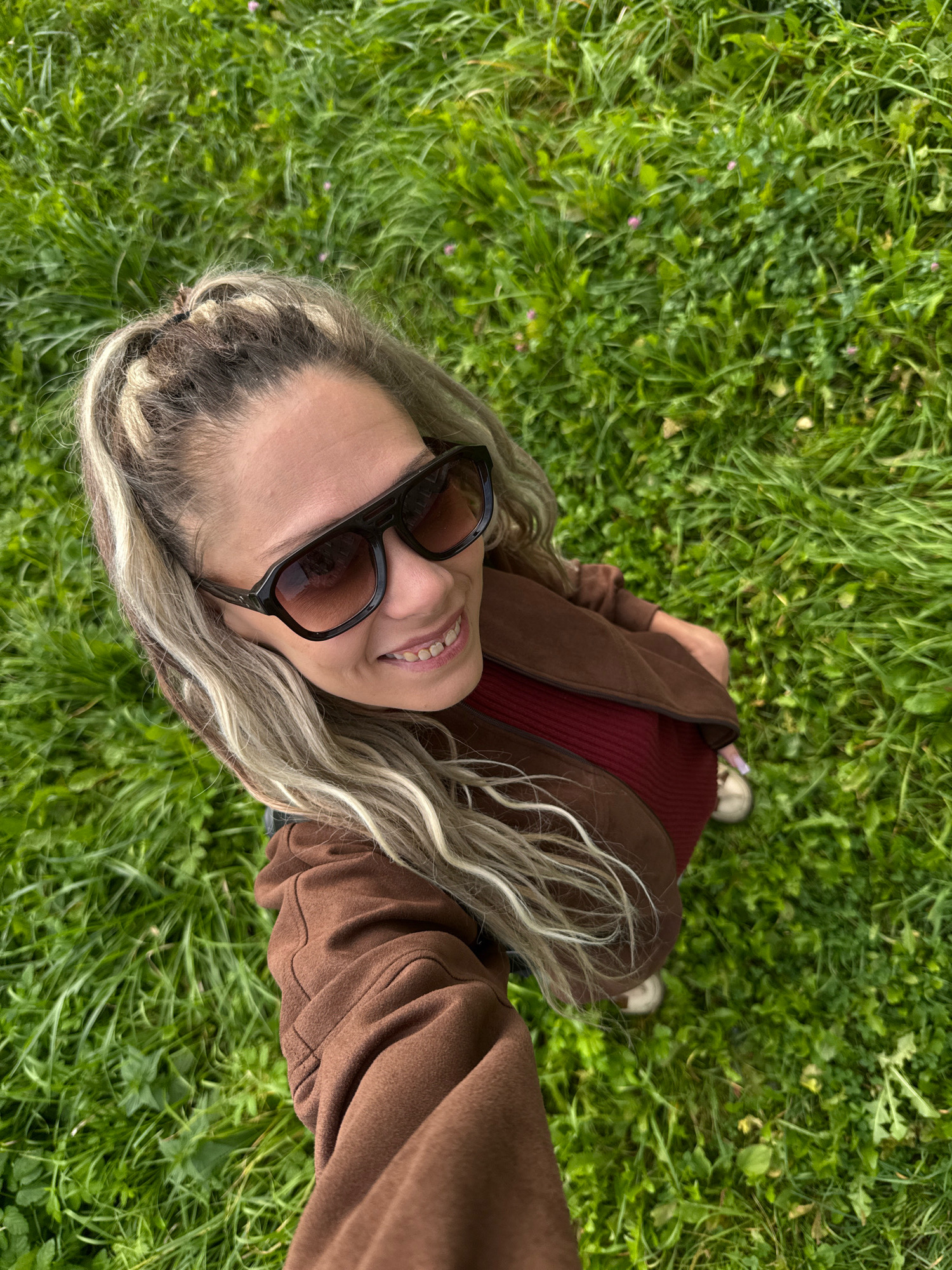 A woman with long, blonde hair, styled in a braid and loose strands, is captured from a high angle in a field of green grass. She is wearing large, dark sunglasses and a brown jacket over a red ribbed top. The woman is smiling, and her left hand appears to be holding a phone, suggesting she is taking a selfie. The grass is lush and appears to be a natural outdoor setting. The specific location in Zhukovo, Russia, cannot be determined from the visual information provided.