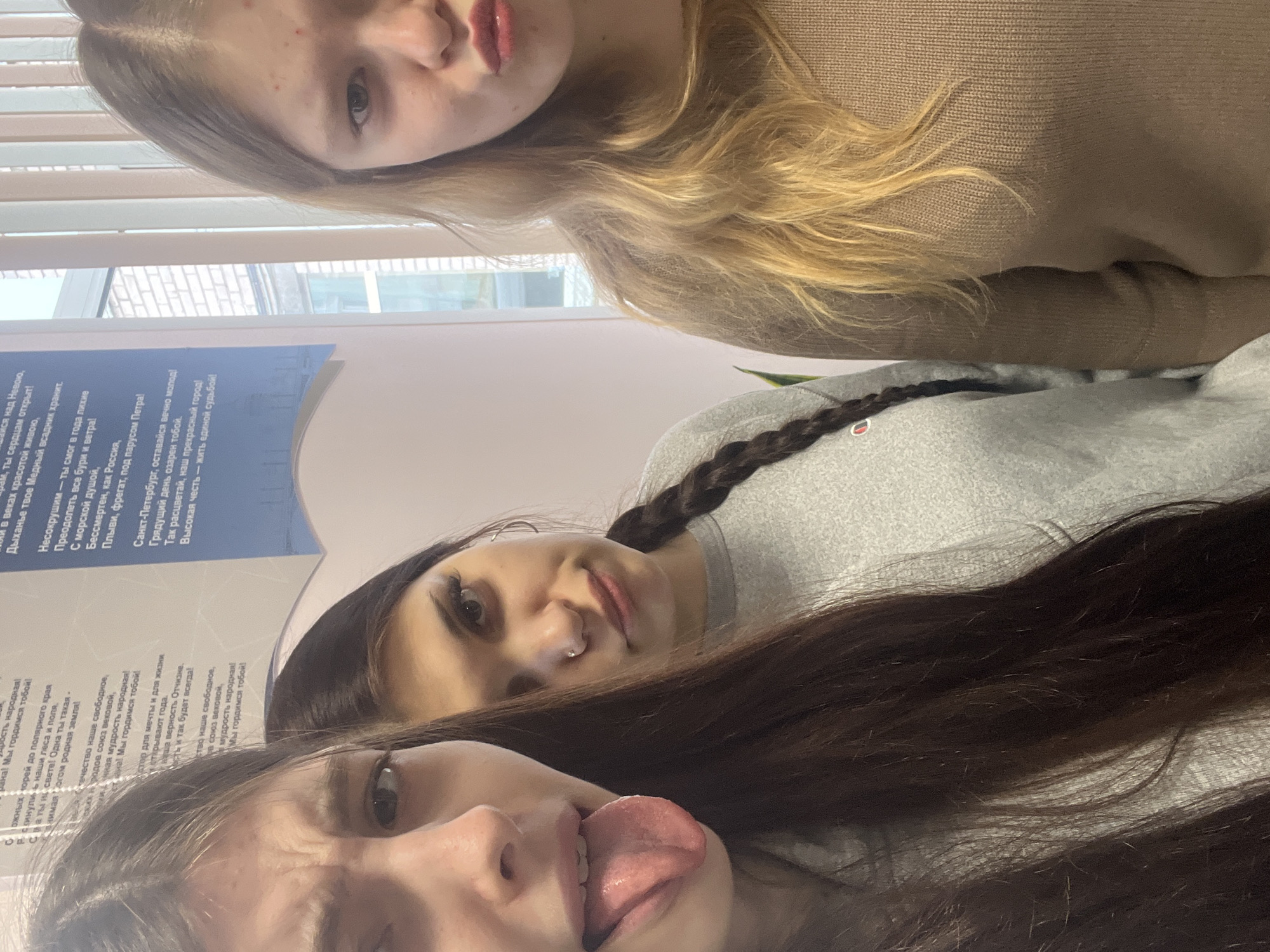 Three young women are in a room, likely a classroom, based on the presence of a wall-mounted poster with Russian text. The poster appears to be about Saint Petersburg, Russia, mentioning historical figures and characteristics of the city. One woman sticks out her tongue, another has a nose piercing and braided hair, and the third is blond. The lighting is bright and appears to be natural.