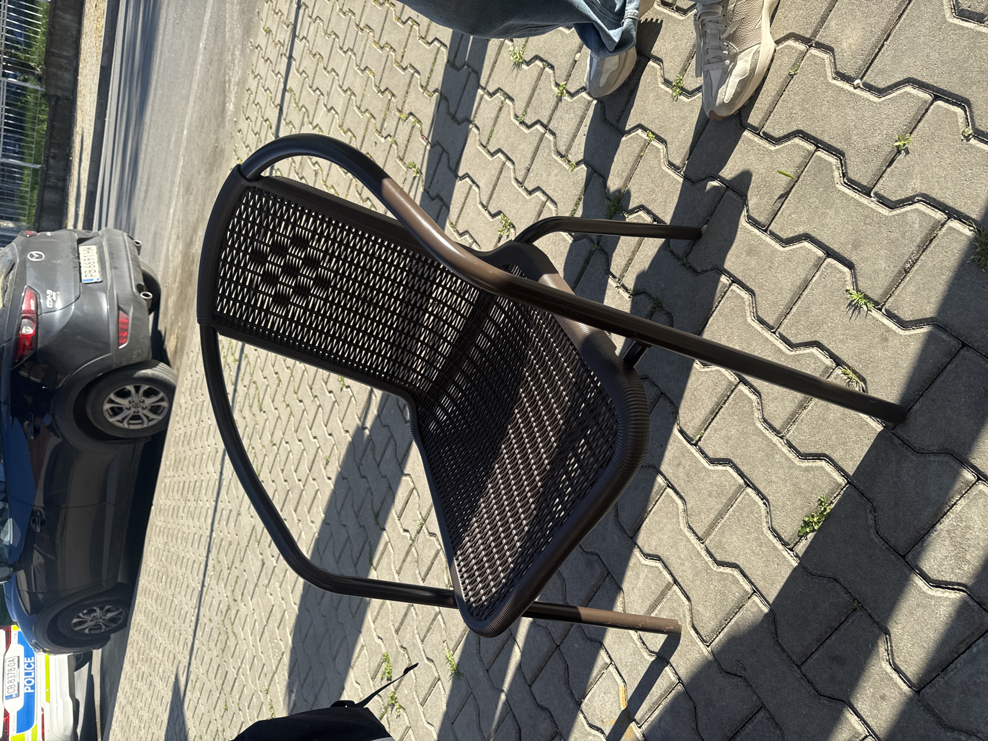 The image shows a brown metal chair with a woven seat and backrest standing on a paved surface made of interlocking gray stones. There is a person in jeans and sneakers standing nearby. In the background, there is a gray Mazda CX-3 car, license plate PB6691HM, and a police car, license plate CB8376OA. The location appears to be a city street, possibly in Unknown, Unknown.