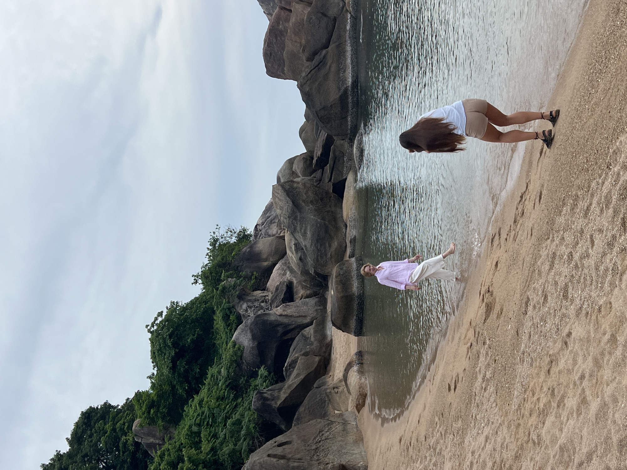 A serene beach scene unfolds at Baan Chaweng Noi, Thailand, under a cloudy sky. The foreground features a sandy beach with gentle waves lapping at the shore. In the middle ground, two women are present. One woman, on the left, is walking in the shallow water, her legs visible as she splashes. She is wearing a light pink long-sleeved shirt and white pants. The second woman, on the right, is facing away from the camera, appearing to take a photo. She is dressed in a white t-shirt and tan shorts, with her long, dark hair cascading down her back. The background is dominated by large, smooth boulders and lush green foliage, characteristic of the island's natural beauty. The water is calm and reflects the muted light of the overcast sky.