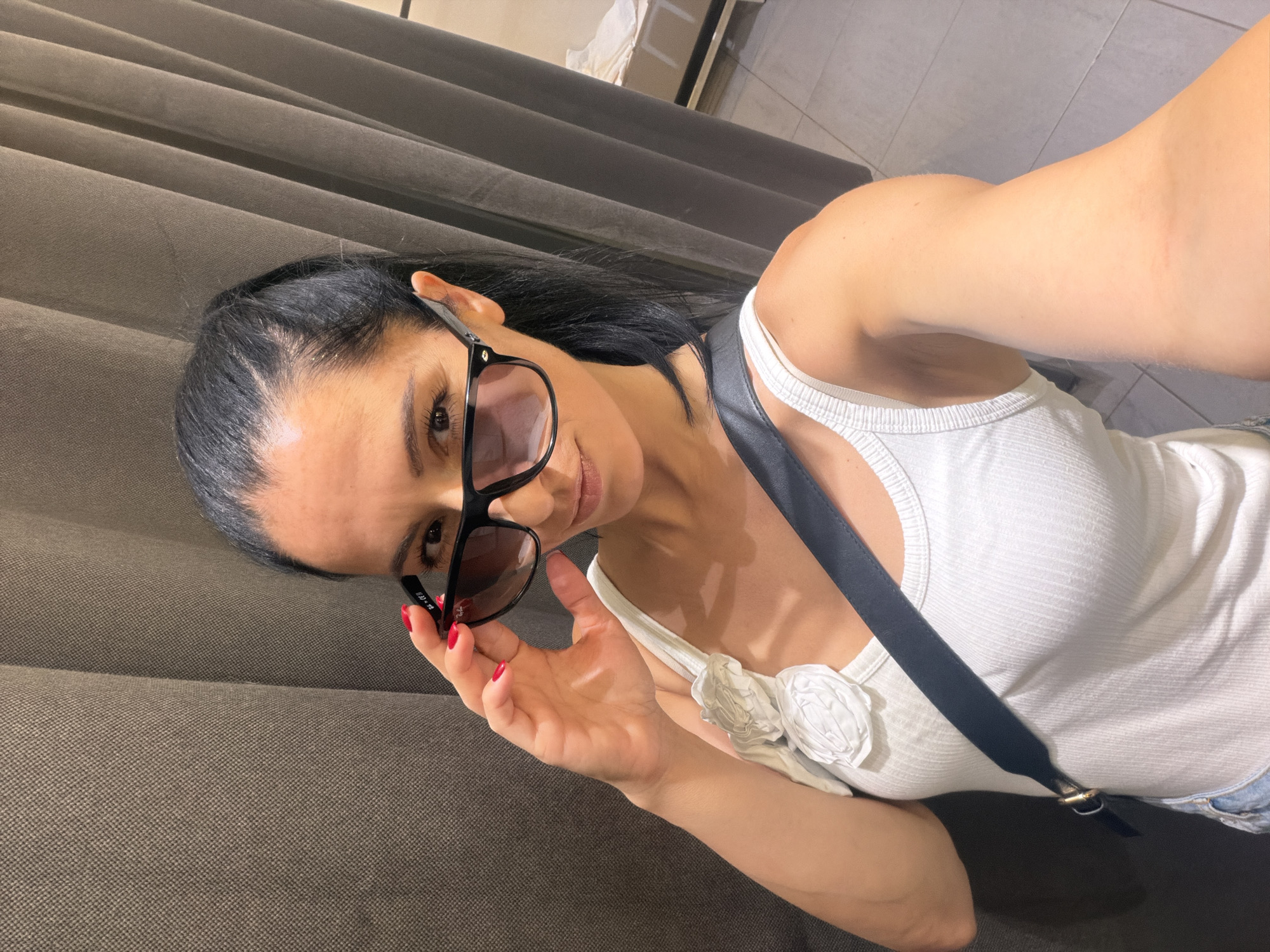 In an indoor setting, possibly a fitting room, a woman with dark hair pulled back in a ponytail is taking a selfie. She is wearing a white tank top with a floral embellishment on the left strap, a black crossbody strap, and sunglasses. Her left hand is raised, adjusting the sunglasses. The background consists of dark, textured curtains. A shelf with white fabric is visible in the upper right corner. The lighting is bright. The image quality is high. The location in the city of Pleven, Bulgaria, is not discernible from the image.