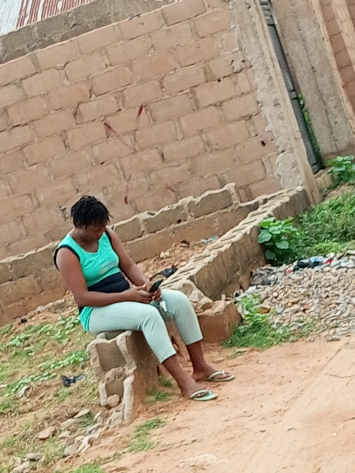A young Black woman sits on a concrete block outdoors, looking down at her smartphone. She is wearing a teal tank top with a black band across the chest and light blue leggings, along with white flip-flops. She has dark, short, braided hair. The background is a rough, tan brick wall. In front of the woman, the ground is sandy with scattered rocks and patches of green grass. To the right of the woman, there is a pile of rubble and more green vegetation. The scene appears to be in an undeveloped or construction area within Tempure, Nigeria.