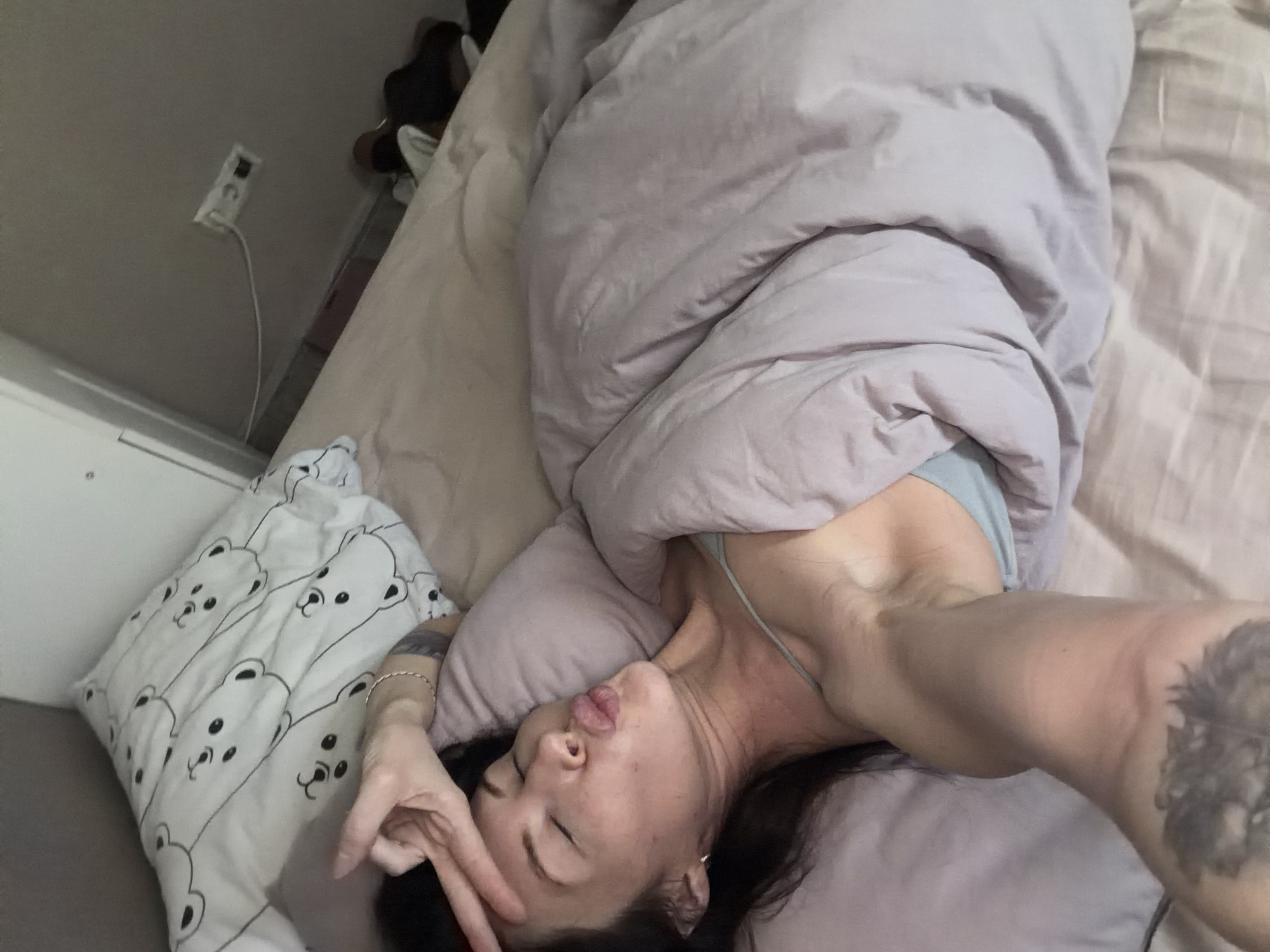 A woman lies in a bed covered with a light-colored duvet and a pillow patterned with cartoon bear faces. She is partially covered by a lavender-colored blanket, exposing one arm with a visible tattoo. Her head is tilted, and she appears to be touching her forehead with her hand. The background includes a wall with an electrical outlet. The specific location in the city of Perm, Russia cannot be determined from this image.