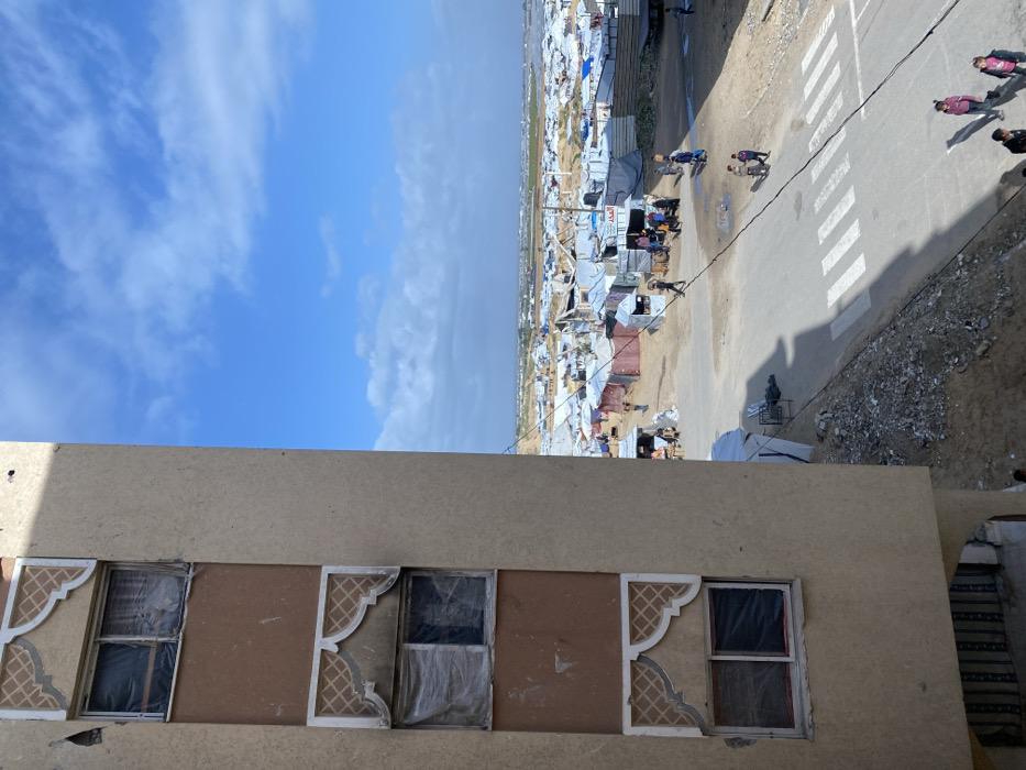 An elevated, high-angle view captures a scene in Khan Yunis, Palestinian Territory. The left side of the frame is dominated by a segment of a multi-story building, constructed with light tan stucco and featuring recessed rectangular panels in a darker brown. Visible on this structure are three pairs of windows, each adorned with an arched, decorative patterned frame above. The windows appear damaged or provisionally covered with clear plastic sheeting and dark fabric.
Below the building, a paved road with a white pedestrian crosswalk cuts diagonally across the midground. Debris and rubble are visible along the road's unpaved edges. Multiple individuals are present: a group of adults and children walk on the paved street, while others are distributed throughout the sprawling encampment in the background.
The background reveals an extensive, densely packed makeshift settlement comprising numerous tents, tarpaulin shelters, and rudimentary structures stretching to the horizon. Many structures appear to be residential, while some larger, more organized clusters, possibly stalls or shops, show gatherings of people. Overhead wires crisscross the air above the encampment and street. Beyond the immediate settlement, the landscape transitions to open, undeveloped terrain under a partly cloudy blue sky.