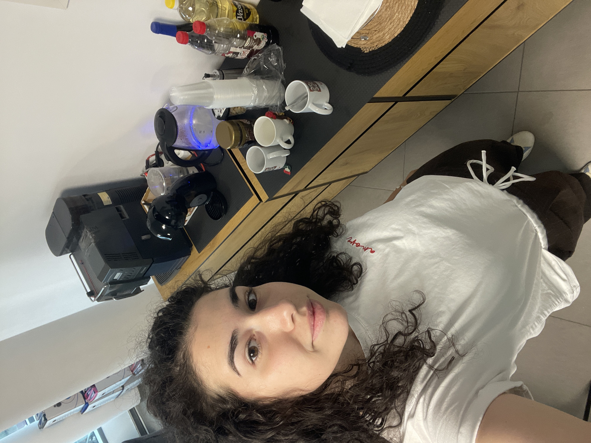 The image depicts an indoor scene with a woman in the foreground. The woman has curly dark hair, is wearing a white t-shirt, and is looking at the camera. Behind her is a table with various items on it. These include bottles of oil and other liquids, disposable cups, coffee mugs, a kettle illuminated with a blue light, and a coffee machine. Part of a woven placemat is visible on the table as well. The floor appears to be tiled. The scene appears to be taken in a kitchen or break room environment. There is no indication of Sofia, Bulgaria.