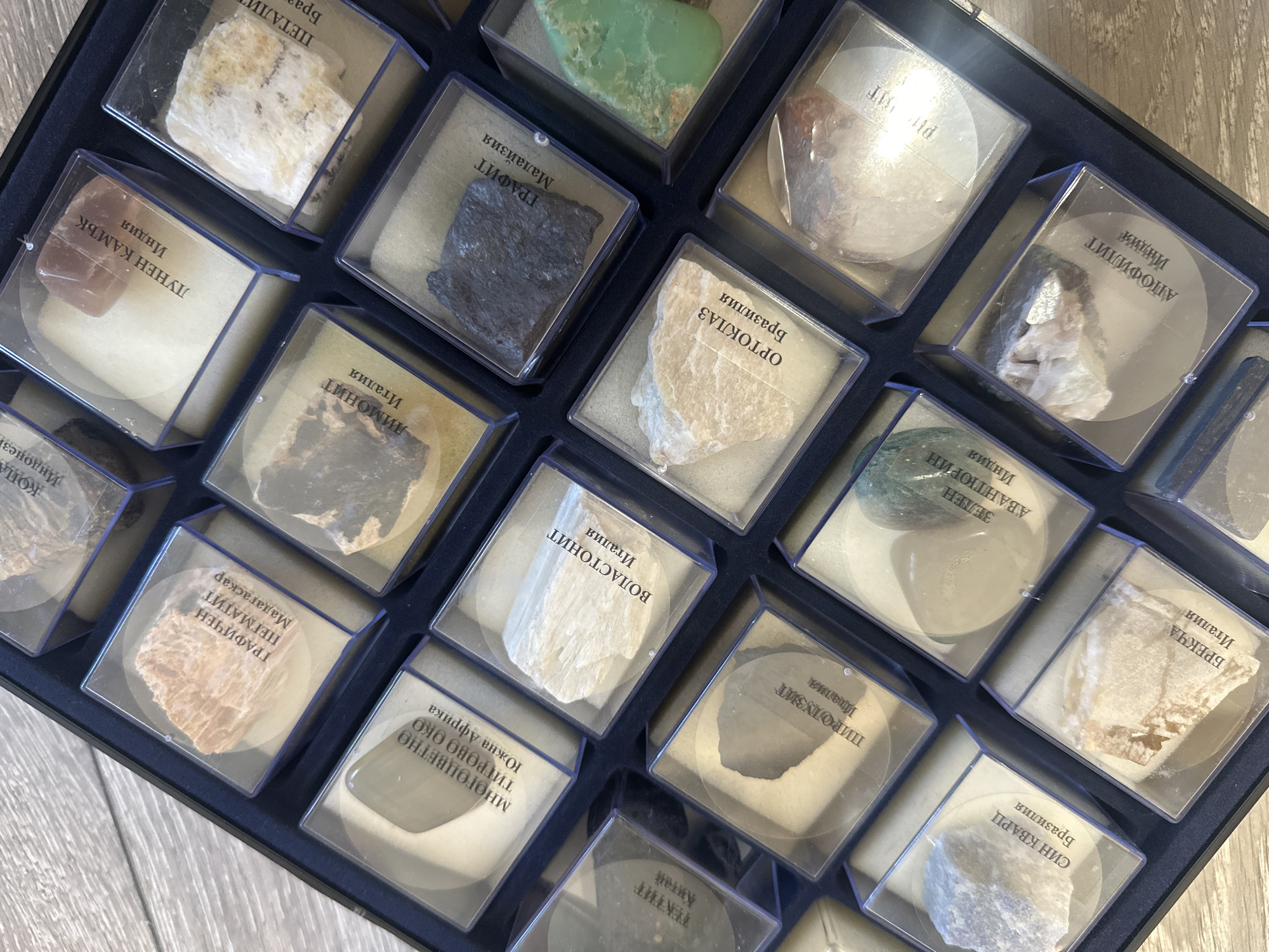 The image shows a collection of various mineral and rock specimens displayed in individual, clear plastic containers arranged in a dark blue tray. Each container features a label with text in Cyrillic, presumably identifying the specimen along with its origin. The specimens include diverse colors and textures, with examples such as a black graphite sample, a green aventurine, a reddish-brown "lunen kamuk" (moonstone), and a blue quartz. The samples are arranged in a grid-like pattern inside the tray. The background appears to be a light wooden surface. There is no information available to discern the location of Sofia, Bulgaria.