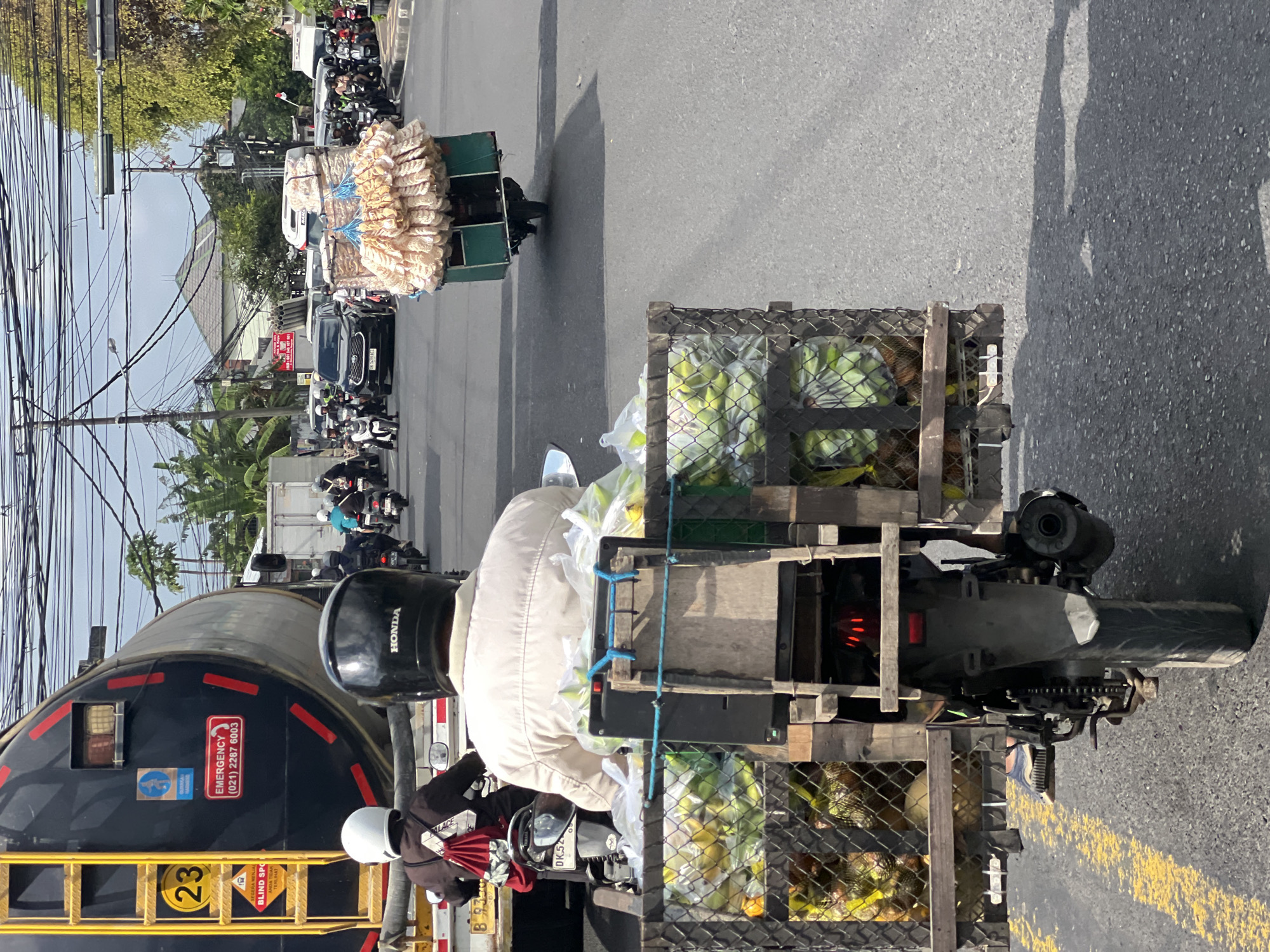 The image captures a street scene in Denpasar, Indonesia, with a focus on a motorcycle carrying a large load of produce. The motorcycle, seen from behind, is traveling on a paved road with yellow lane markings. The rider is wearing a light-colored jacket and a black helmet. Attached to the back of the motorcycle is a wooden and metal cage filled with green fruits, possibly bananas, and other items. To the left, a large black tanker truck with a yellow ladder and safety markings, including an "EMERGENCY" contact number, is visible. Other vehicles, including cars and motorcycles, are present on the road. The background shows buildings, trees, and a complex network of overhead electrical wires, characteristic of urban environments. The sky is partly cloudy. The overall impression is a snapshot of daily life and commerce on the streets of Denpasar.