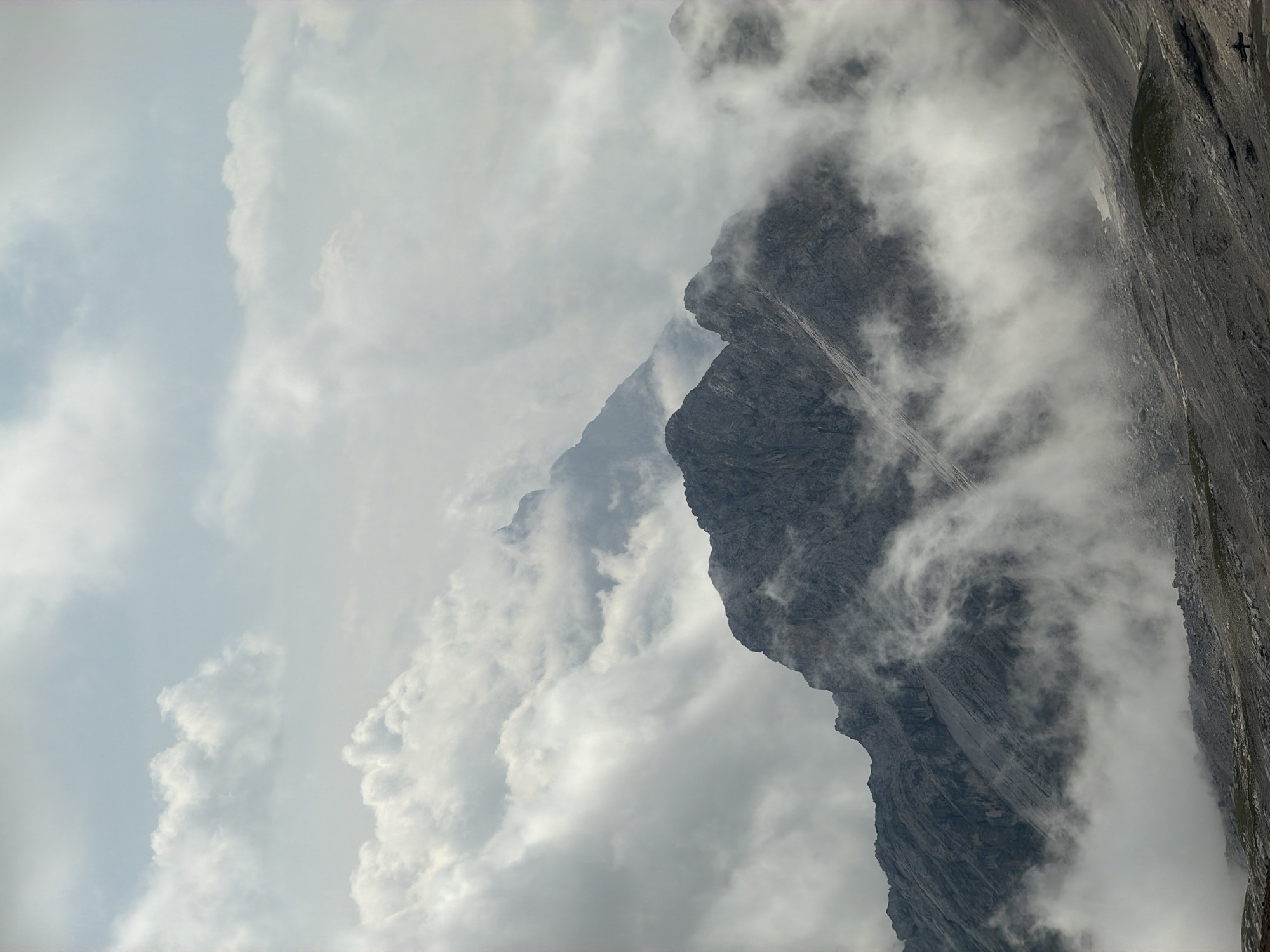 The image shows a dramatic mountain landscape with sharp, rocky peaks partially obscured by swirling clouds and mist. The foreground is dominated by a rugged, gray mountain ridge with visible scree slopes. In the background, additional mountain peaks emerge from the mist, some appearing as sharp silhouettes against the sky. The sky is overcast with patches of bright, luminous clouds interspersed with darker, more diffuse ones. The overall impression is one of a wild and imposing natural environment, possibly taken from a high vantage point. This scene is characteristic of the Bavarian Alps near Garmisch-Partenkirchen, Germany, a region renowned for its majestic mountain ranges and frequent cloud formations at high altitudes. There are no people or identifiable structures visible in the immediate scene.