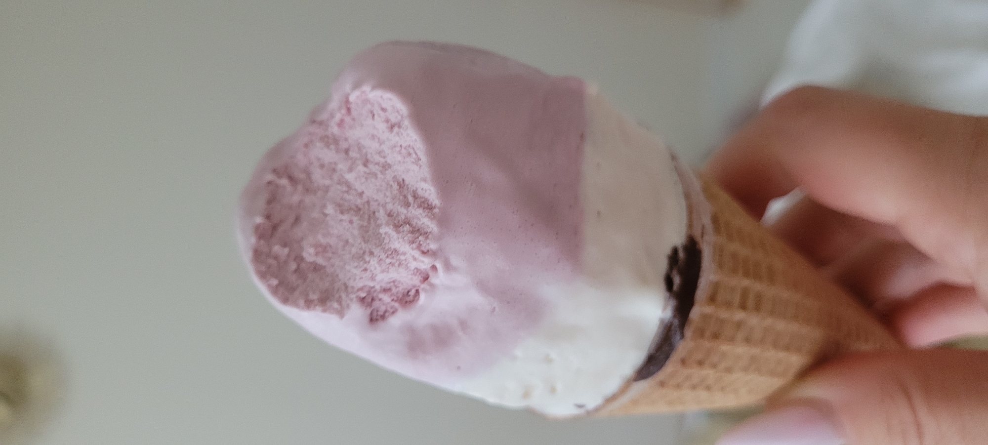 The image shows a close-up of a person's hand holding an ice cream cone. The ice cream has two layers: a white layer on the bottom and a pinkish-purple layer on top. There is a bite taken out of the top layer. The ice cream is in a waffle cone. The background is out of focus and appears to be a light-colored wall. There are no specific landmarks or clues in the image to indicate a location in Riga, Latvia. The image is a close-up shot, so only the hands and the ice cream are clearly visible.