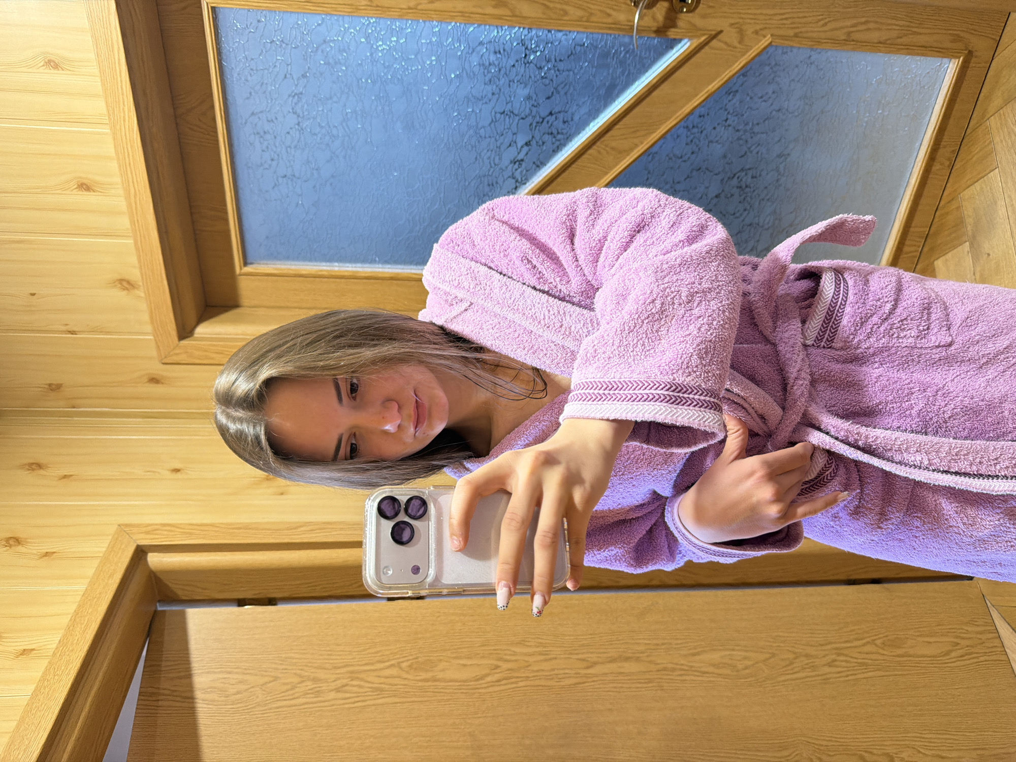 A young woman in a pink bathrobe is taking a selfie with her smartphone. The background shows a wooden door with a frosted glass pane. The scene appears to be indoors, potentially in Buhovo, Bulgaria, although the location cannot be confirmed without additional information.