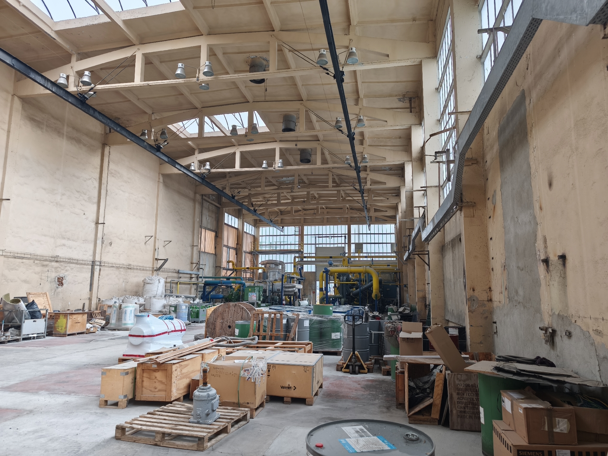This image depicts the interior of a large industrial warehouse or factory floor, illuminated by natural light streaming through numerous windows along the upper walls and skylights in the ceiling, augmented by overhead high-bay lighting fixtures. The space features a robust structural design with exposed concrete beams and trusses, and a dark metal beam spanning the width of the building.
The floor is occupied by various industrial materials and equipment. In the foreground and midground, there are numerous wooden crates and pallets. Some crates hold items wrapped in white plastic, including a large cylindrical object, potentially a machinery component. A gray mechanical part rests on one pallet. Other items include a large wooden spool, green and gray industrial barrels, sacks of material, and cardboard boxes. A pallet jack is visible among the barrels. In the background, an array of industrial machinery with intricate yellow and blue piping, tanks, and what appear to be large engines or compressors is arranged. The walls exhibit signs of age and use, with patched and peeling paint.
No people are present in the scene, suggesting a temporary lull in activity or that the space is set up for storage or machinery installation. The environment appears functional and utilitarian.
Visible text includes brand names "Valmet" and "Siemens" on cardboard boxes. A wooden crate displays detailed text such as "PO 2064-J", "SOL BULGA", "NEW TECHNI", "ACROPOLY", "INDUSTRIA", "CRUTIKO", "DEHENSIONS", and "MADE IN TH". Additional text "77", "A2024", and "Fed" is visible on other packages.
