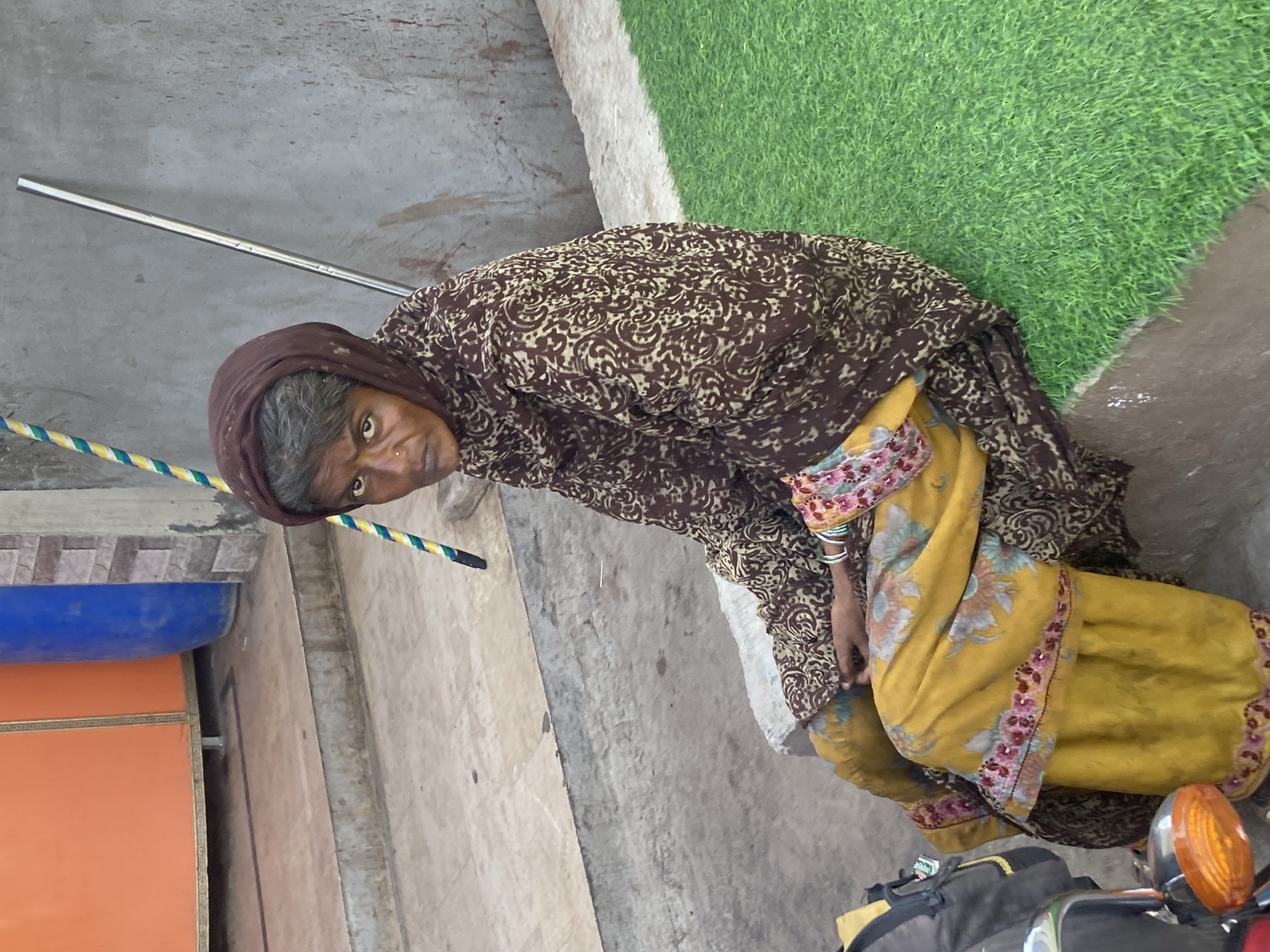An older woman sits on a low platform in Uthal, Pakistan. She is wearing a brown patterned shawl and a yellow salwar kameez with floral accents. To her right is a motorcycle with a visible headlight. To her left is a building with a blue awning. A patch of green artificial grass is next to the platform on which she sits. A metal bar is also visible.