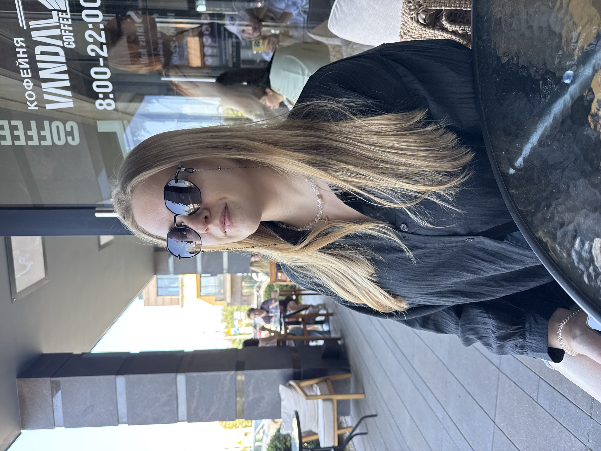 A blonde woman wearing sunglasses and a black collared shirt sits at an outdoor table, looking to her left. She has a beaded necklace on and her hair is long and blonde, falling over her shoulders. The background shows a street with a building that has the words "КОФЕЙНЯ VANDAL COFFEE" and the hours "8:00-22:00" visible on its windows. Other patrons are seated at tables on the patio. The scene is captured in daylight with clear skies.