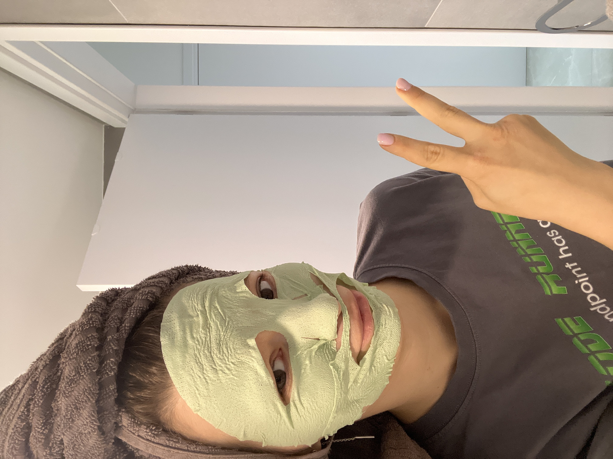 A person wearing a gray shirt and a green face mask is making a peace sign with their right hand. The person also has a brown towel wrapped around their head. The setting appears to be a bathroom with white walls and a white cabinet. The city and location are unknown.