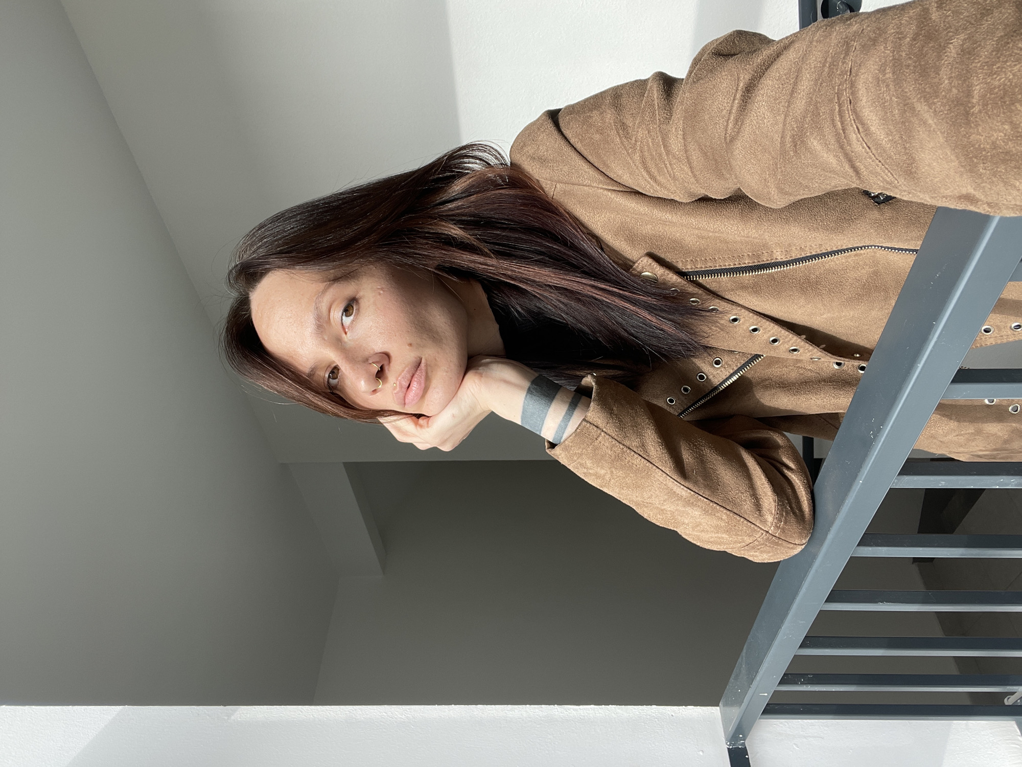 A young woman with long, dark brown hair is captured in a vertical, low-angle shot, looking downwards with a pensive expression. She rests her chin on her left hand, revealing a tattoo of several thick, dark bands on her forearm. Her eyes are light-colored, and she has a small stud and hoop piercing in her nose. She is wearing a brown jacket, appearing to be made of suede or a similar material, featuring a prominent zipper, eyelet details, and a high collar.
The woman is leaning over a grey metal structure, possibly a railing or balustrade, which features horizontal bars. The background consists of light grey walls, with strong, natural daylight streaming in from the upper right, casting distinct shadows on the surfaces. This suggests a bright daytime setting, possibly indoors within a modern building. The scene depicts a quiet, contemplative moment in Izhevsk, Russia. No visible text is present.